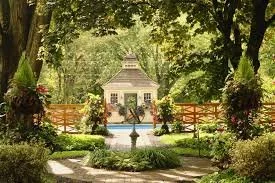 A small white gazebo surrounded by lush green trees and flower pots, with a wooden fence and pathway leading to it. This venue is The Cypress House in New Columbia, PA near Lewisburg, PA.