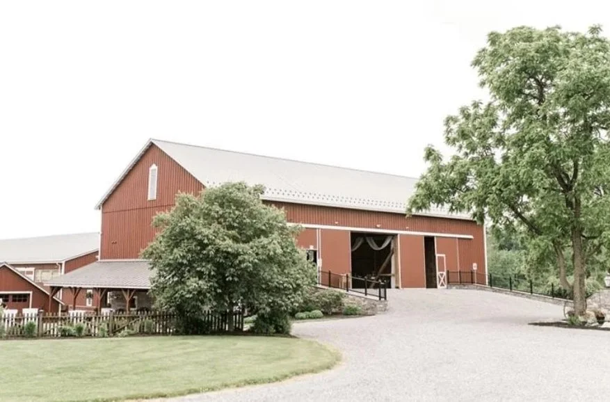 Red barn with white roof, Called the Barn at Greystone, surrounded by green trees and landscaped yard, gravel driveway in front. Located in Turbotville, PA, near Lewisburg, PA.