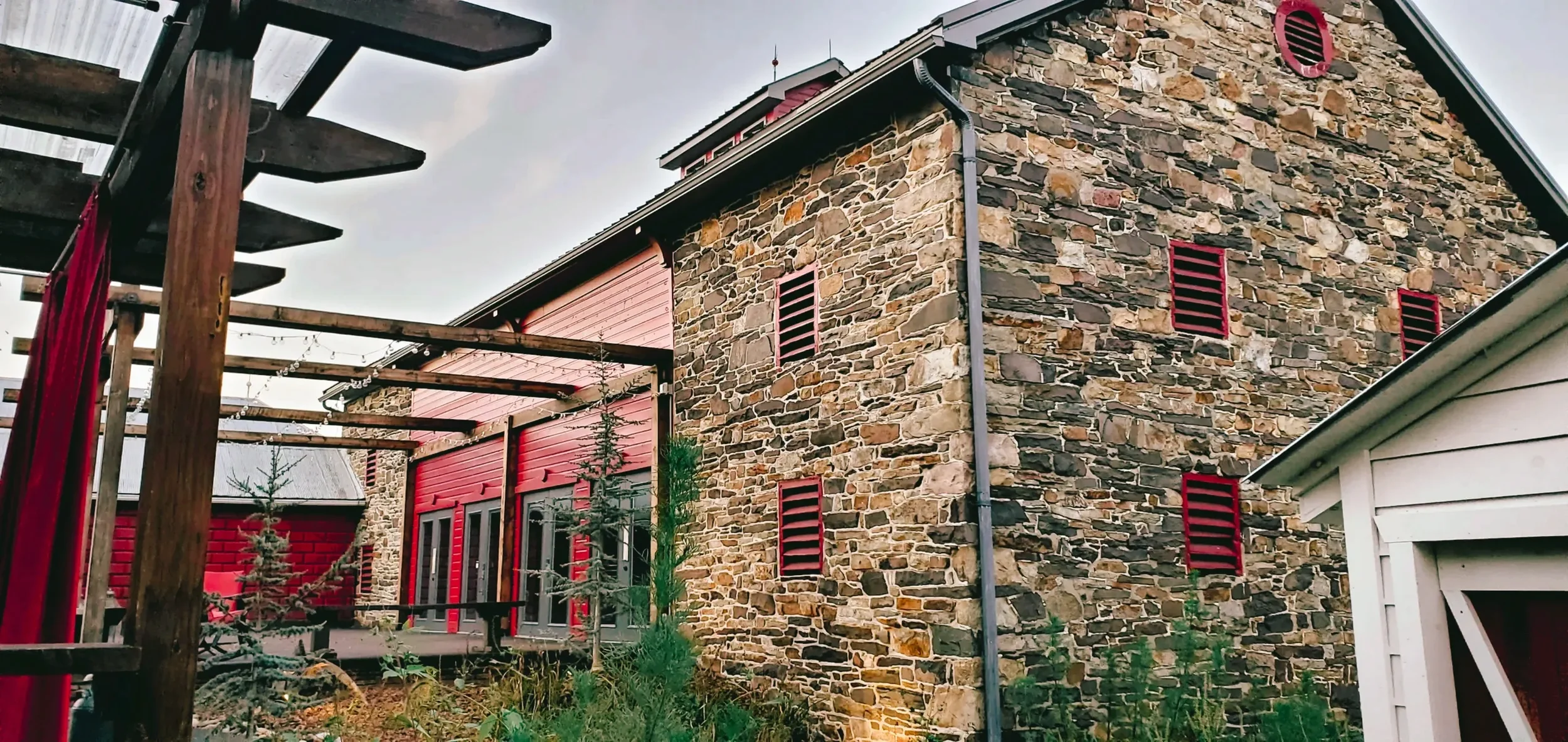A stone house with red window shutters and a metal roof. To the left, there is a wooden pergola with string lights and small trees. A barn venue called Stone Barn Farm in Selinsgrove, PA near Lewisburg, PA where Plate & Platter caters.