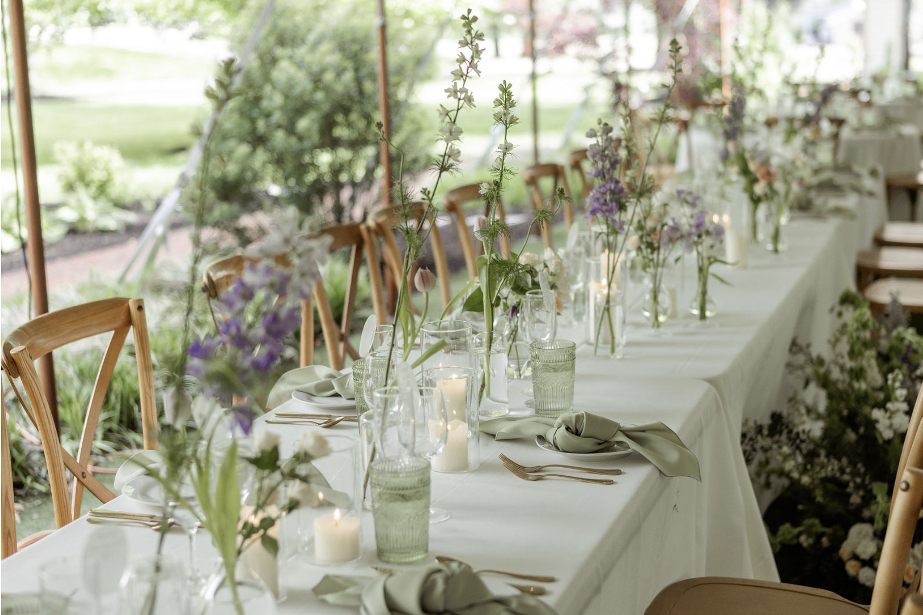 Long banquet table set for a wedding reception with white tablecloths, floral centerpieces in tall vases, lit candles, and place settings with gold utensils and green napkins. Wooden chairs surround the table inside a tent with a view of a garden outside.