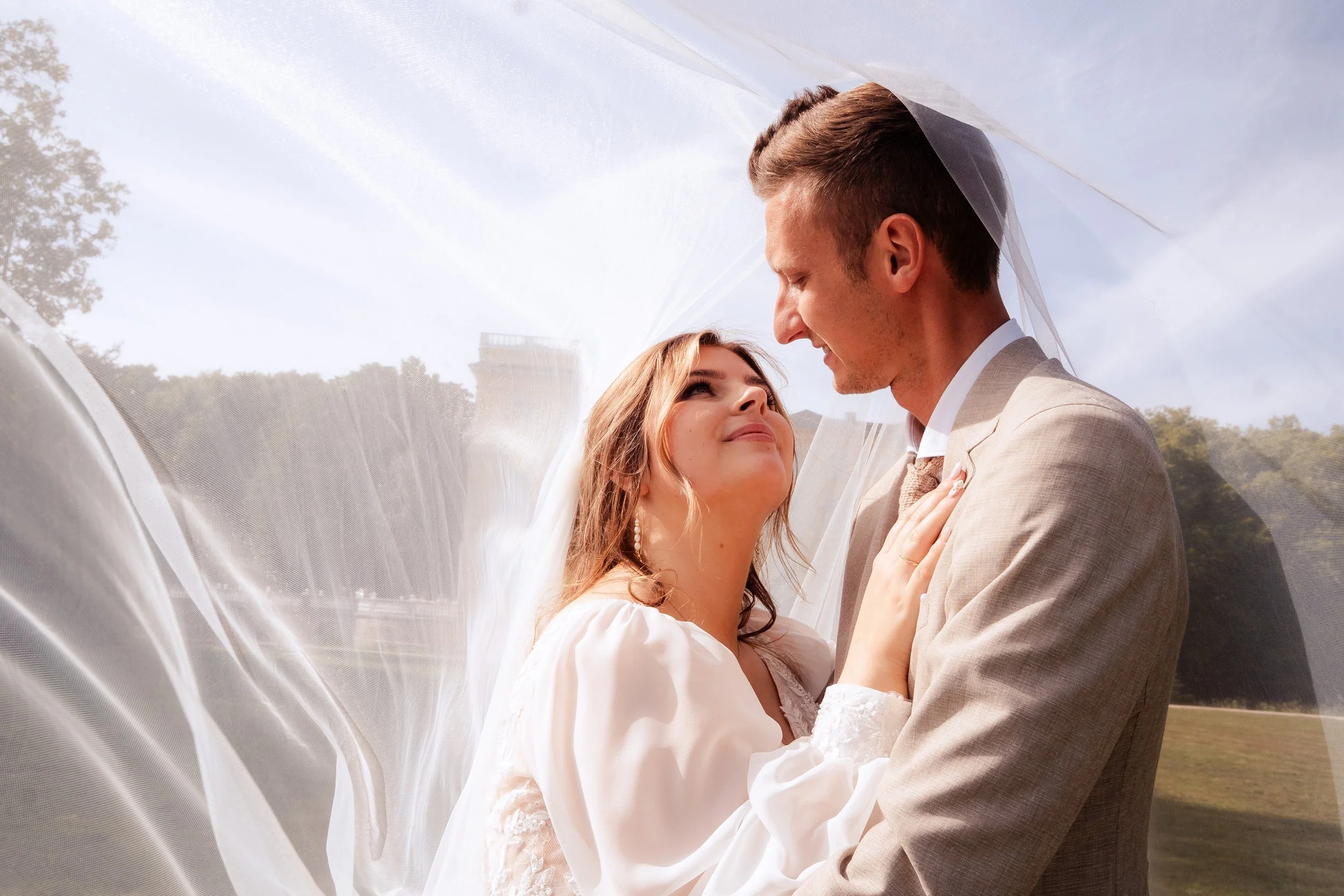 Bruidal stel dat elkaar liefdevol aankijkt onder een witte sluier buiten.