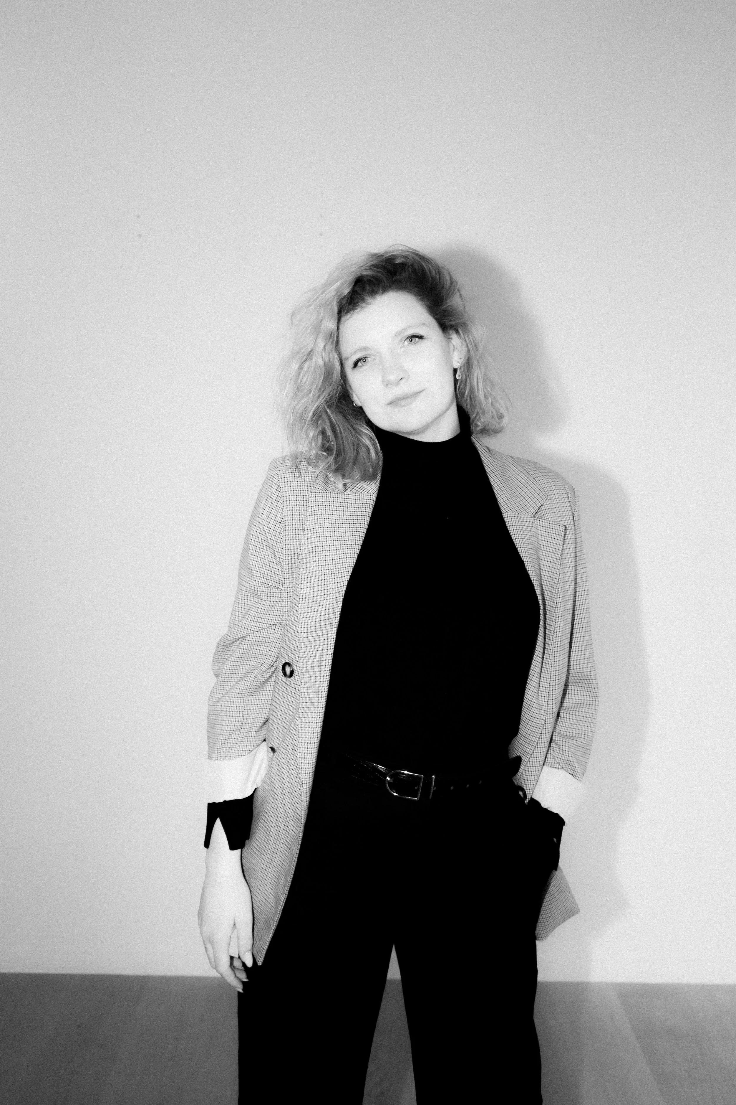 Zwarte en witte portretfoto van een vrouw met schouderlange golvende haar, gekleed in een zwarte top en een blazer, staand tegen een effen muur.