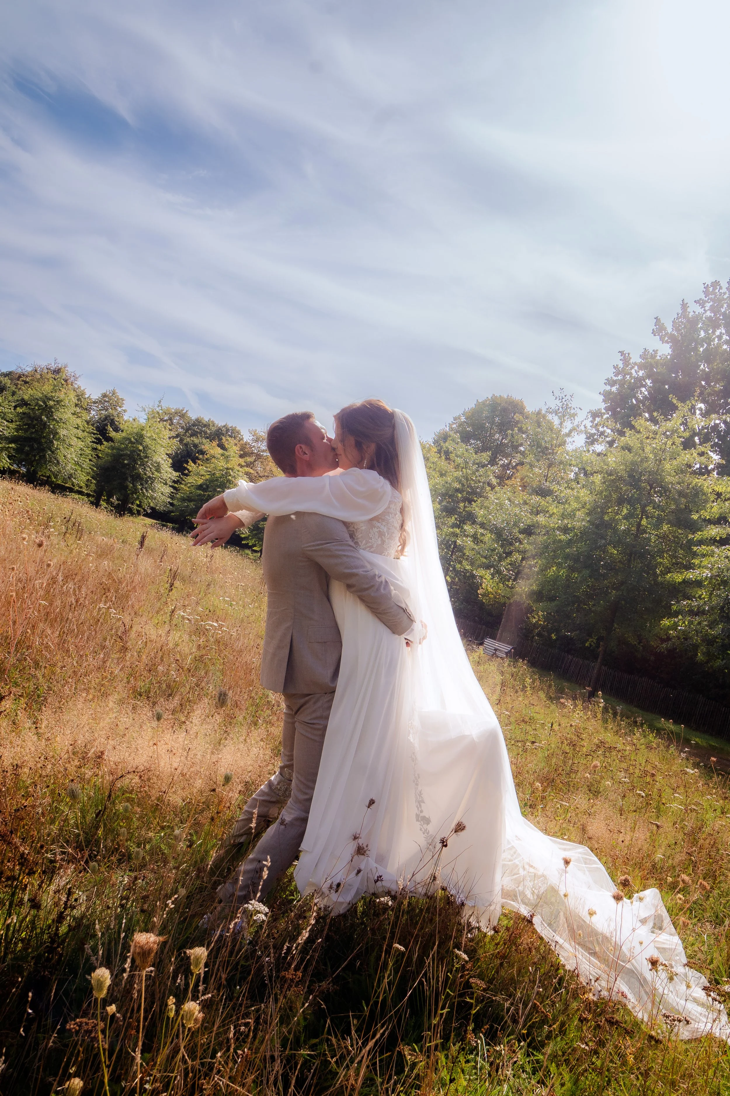 Een bruid en bruidegom die elkaar in een veld kussen, met groene bomen en een blauw luchtruim op de achtergrond.
