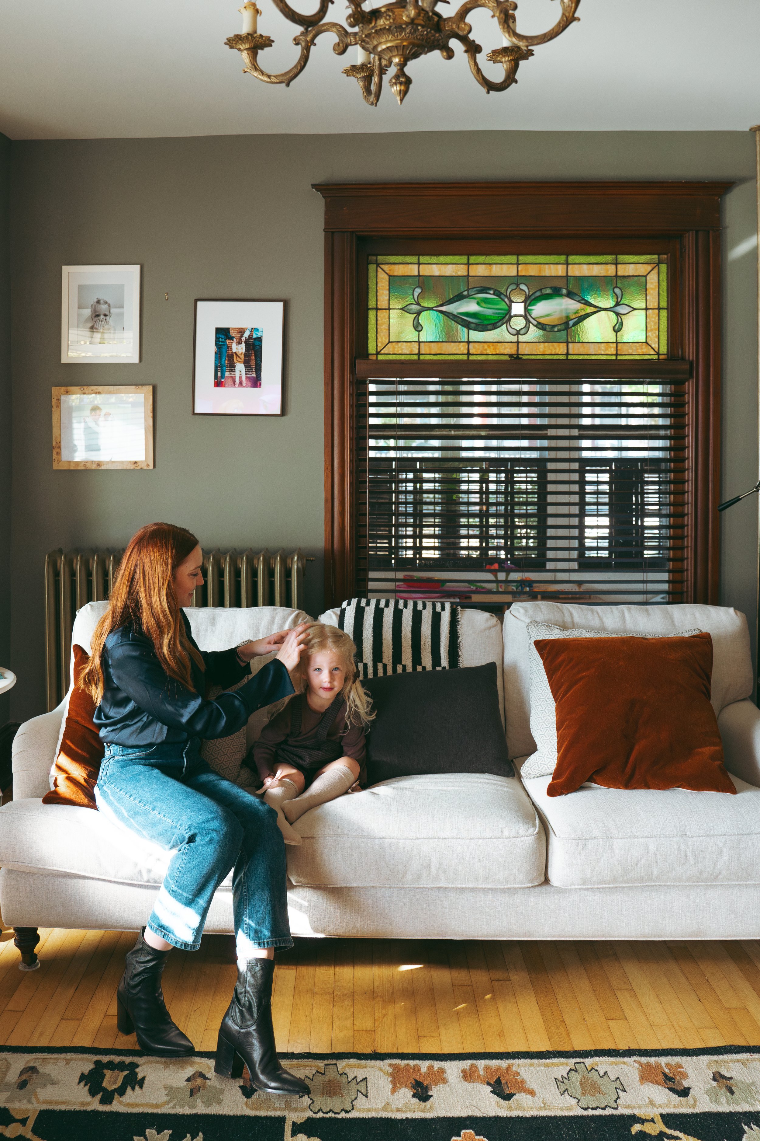 A woman with long red hair wearing a black jacket, blue jeans, and black heeled boots, is sitting on a cream sofa, styling a young girl’s blonde hair. The girl is sitting on the sofa in a black dress, beige tights, and beige shoes. Behind them is a d