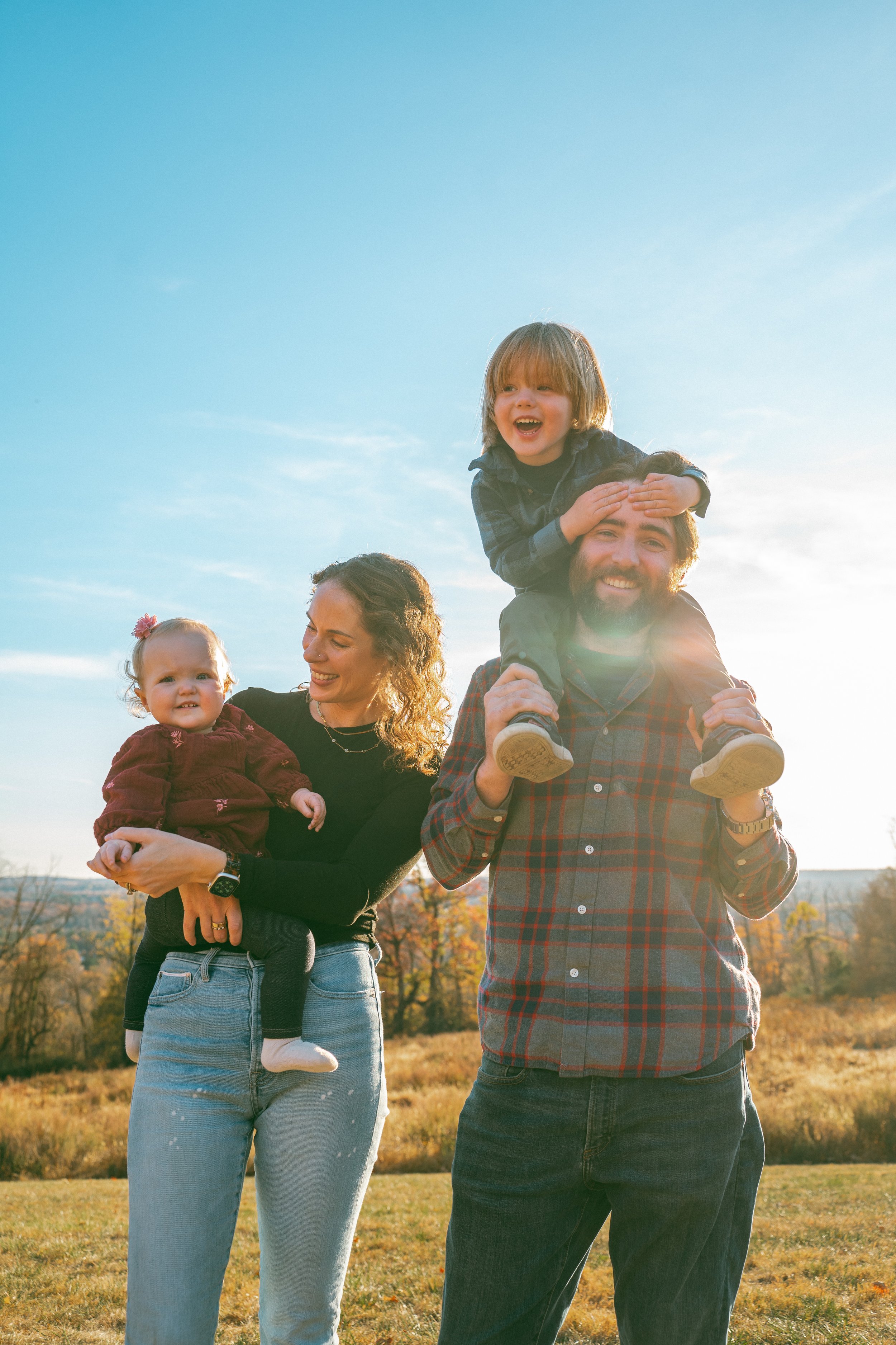 A diverse family of four enjoying outdoors during sunset, with the father carrying a smiling boy on his shoulders, the mother holding a girl, and trees in the background.