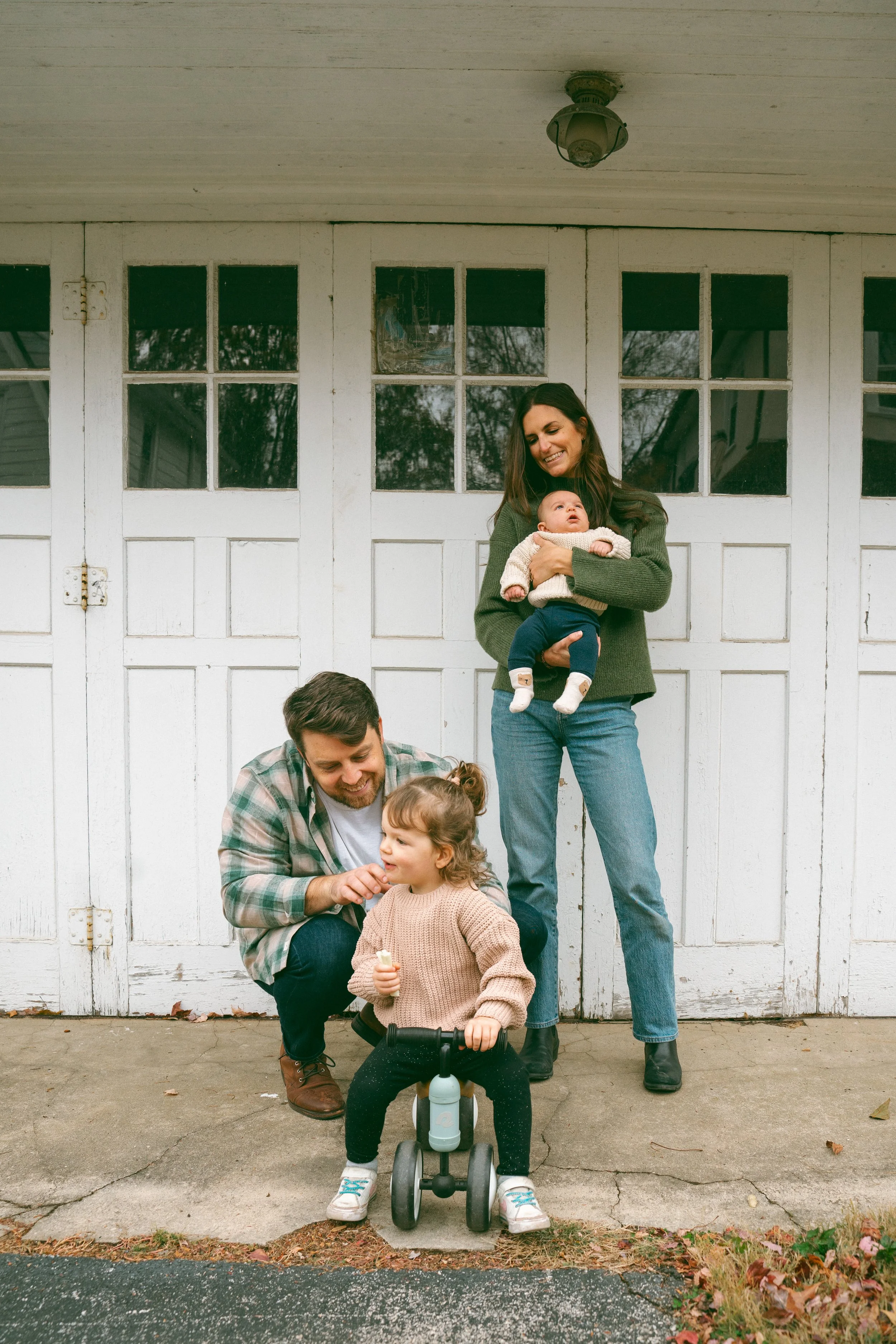 A woman holding a baby while smiling standing in front of a white garage door outdoors. A man crouches down, playing with a young girl on a tricycle, in front of her. Family enjoying time together on a paved driveway with fallen leaves.