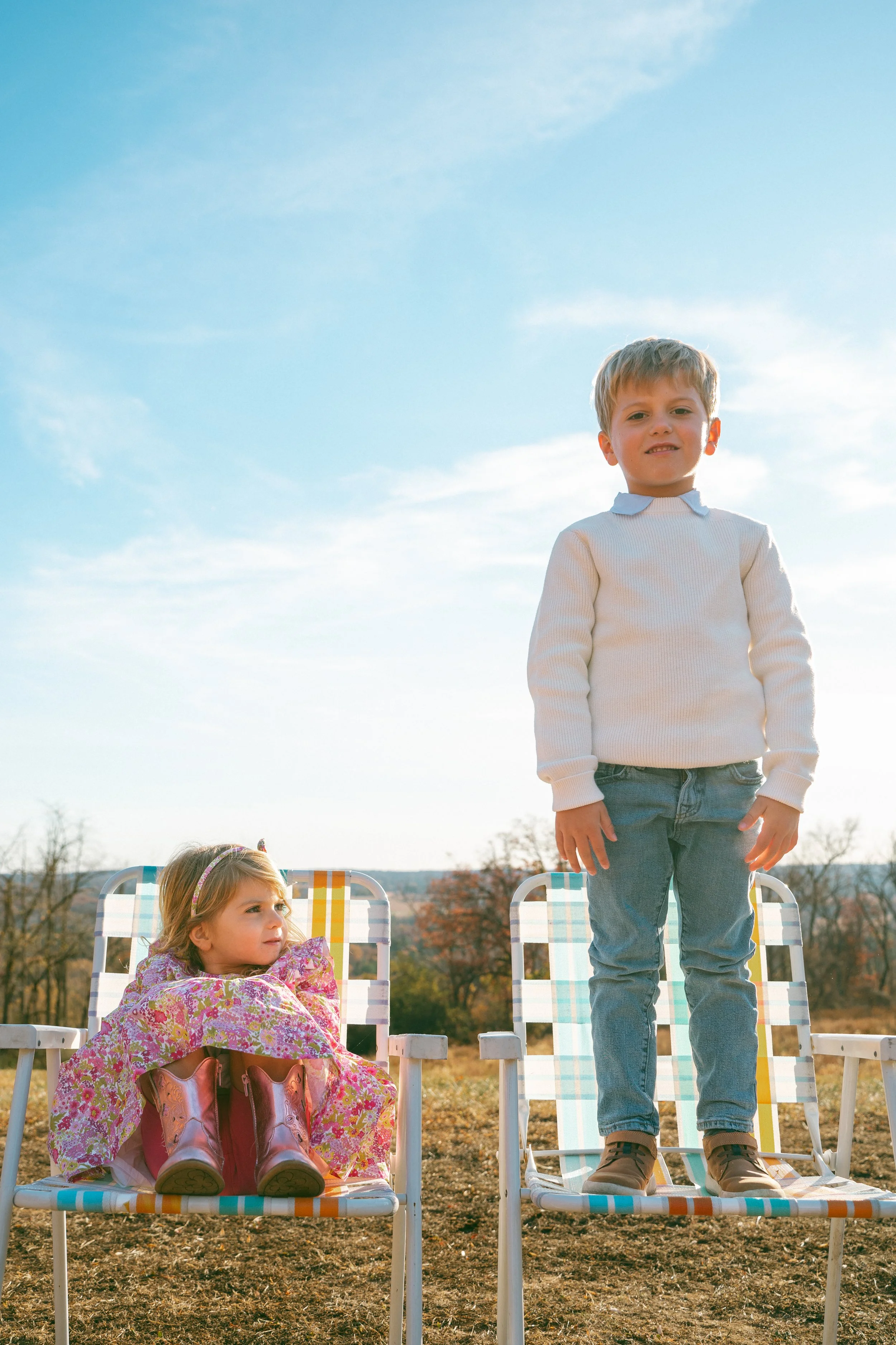 A young boy standing on a colorful lawn chair outdoors during daytime with a girl sitting in a similar chair beside him. The boy is dressed in a light sweater and jeans, while the girl wears a patterned dress and cowboy boots, and is wrapped in a flo