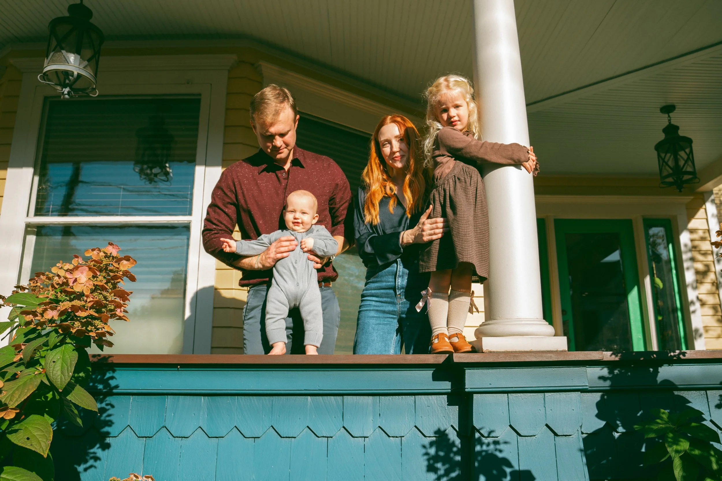 Family standing on porch of yellow house with white trim, a man holding a young baby, a red-haired woman, and a young girl with blonde hair leaning on a white porch column, with plants in the foreground.