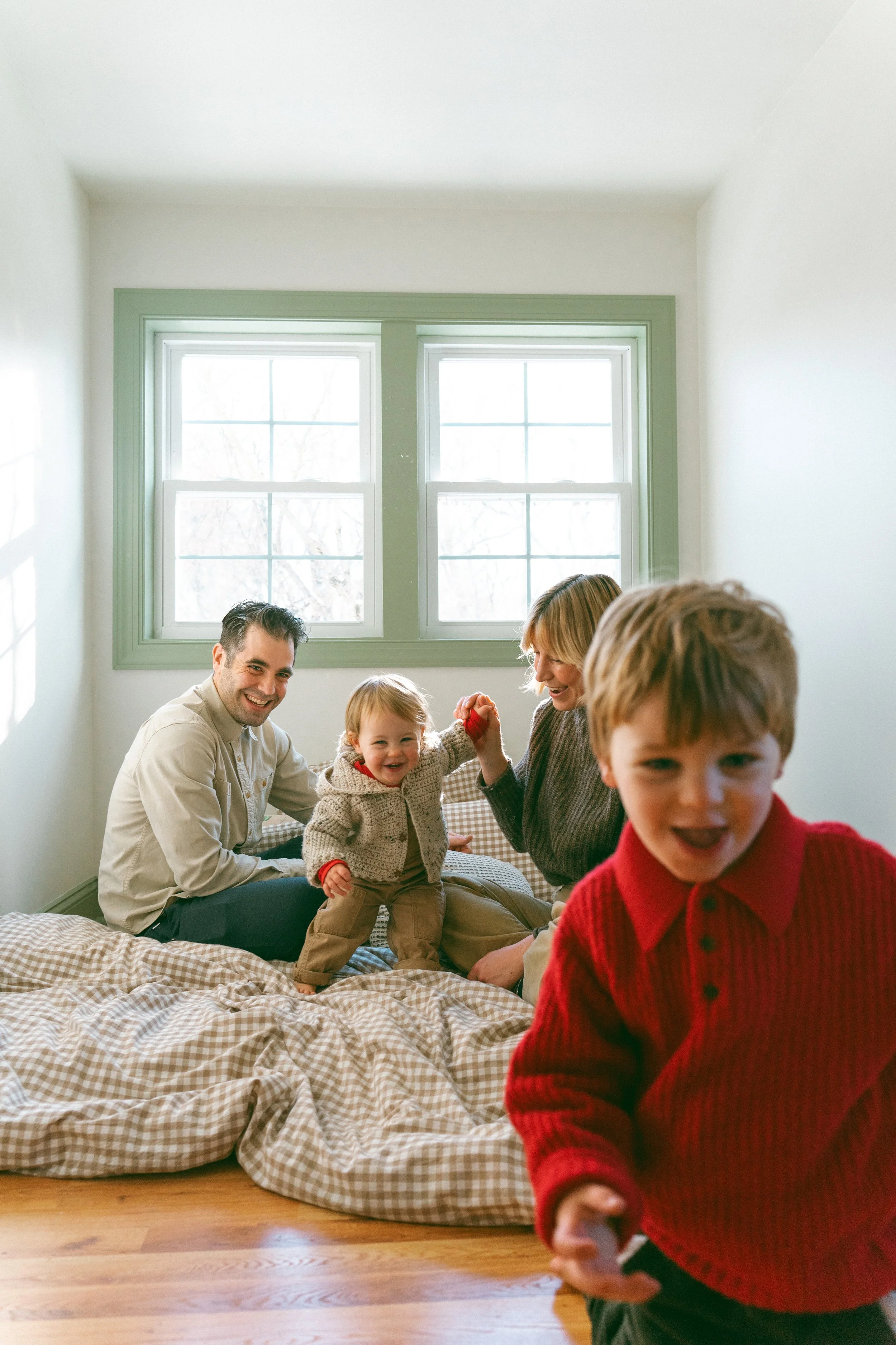 A family of five enjoying time together in a cozy room with a large window and checkered bedding. The adults and children are smiling, with one child in a red sweater in the foreground, and a toddler, a man, and a woman sitting on the bed.