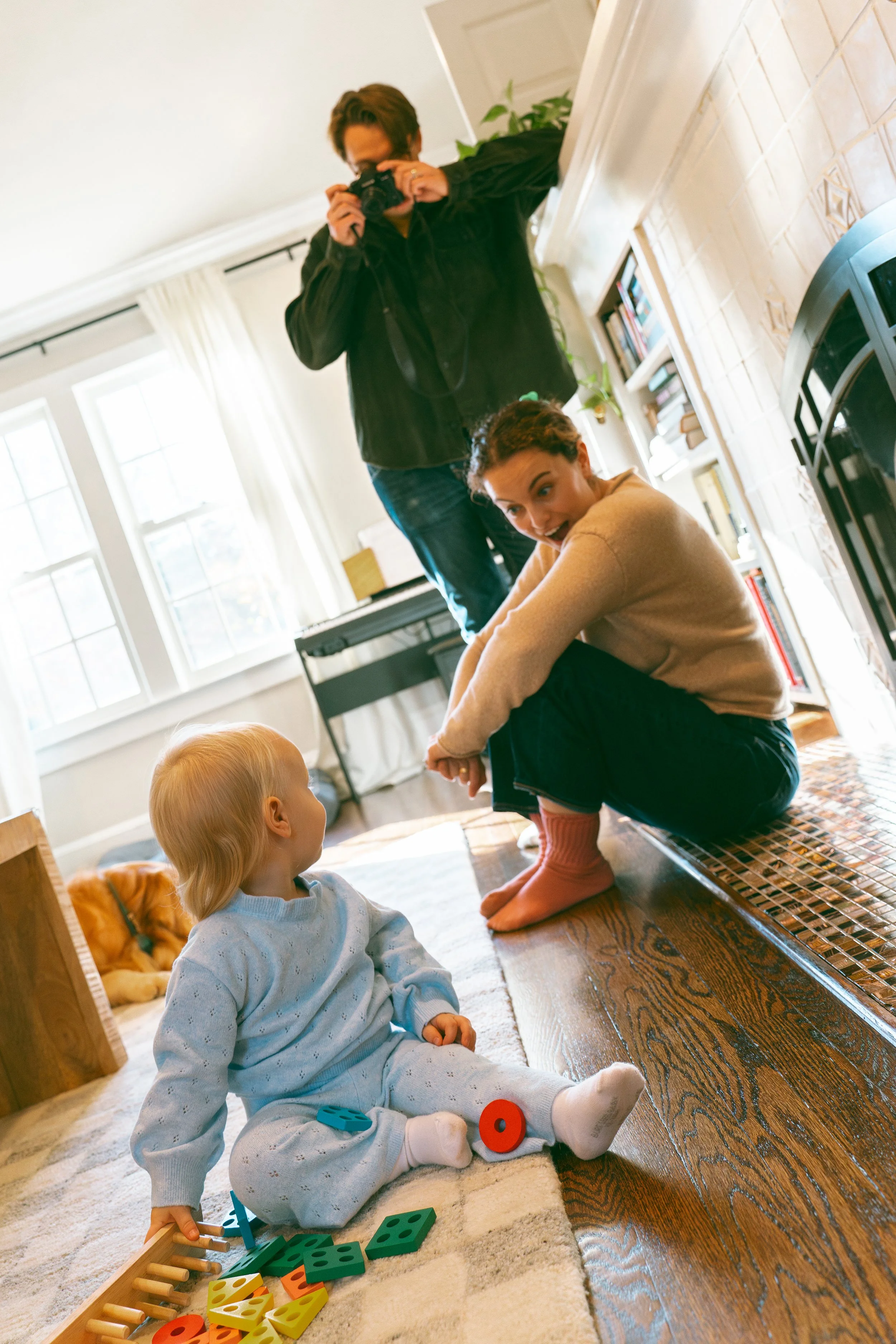 A woman with curly hair sitting on the floor playing with children, one child sitting on the floor looking at her, another child standing and taking a picture, in a cozy living room with large windows, bookshelves, and a fireplace.