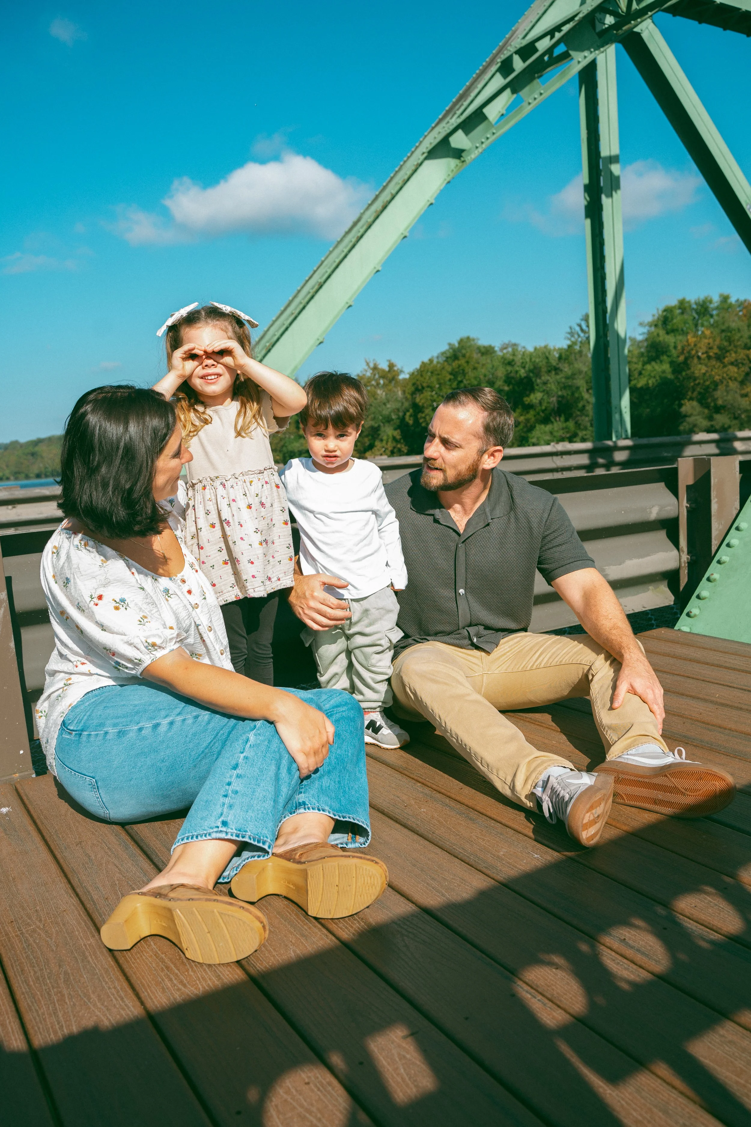 A family of four, including a woman, a man, and two children, sitting on a wooden deck with a green bridge and trees in the background, enjoying a sunny day.