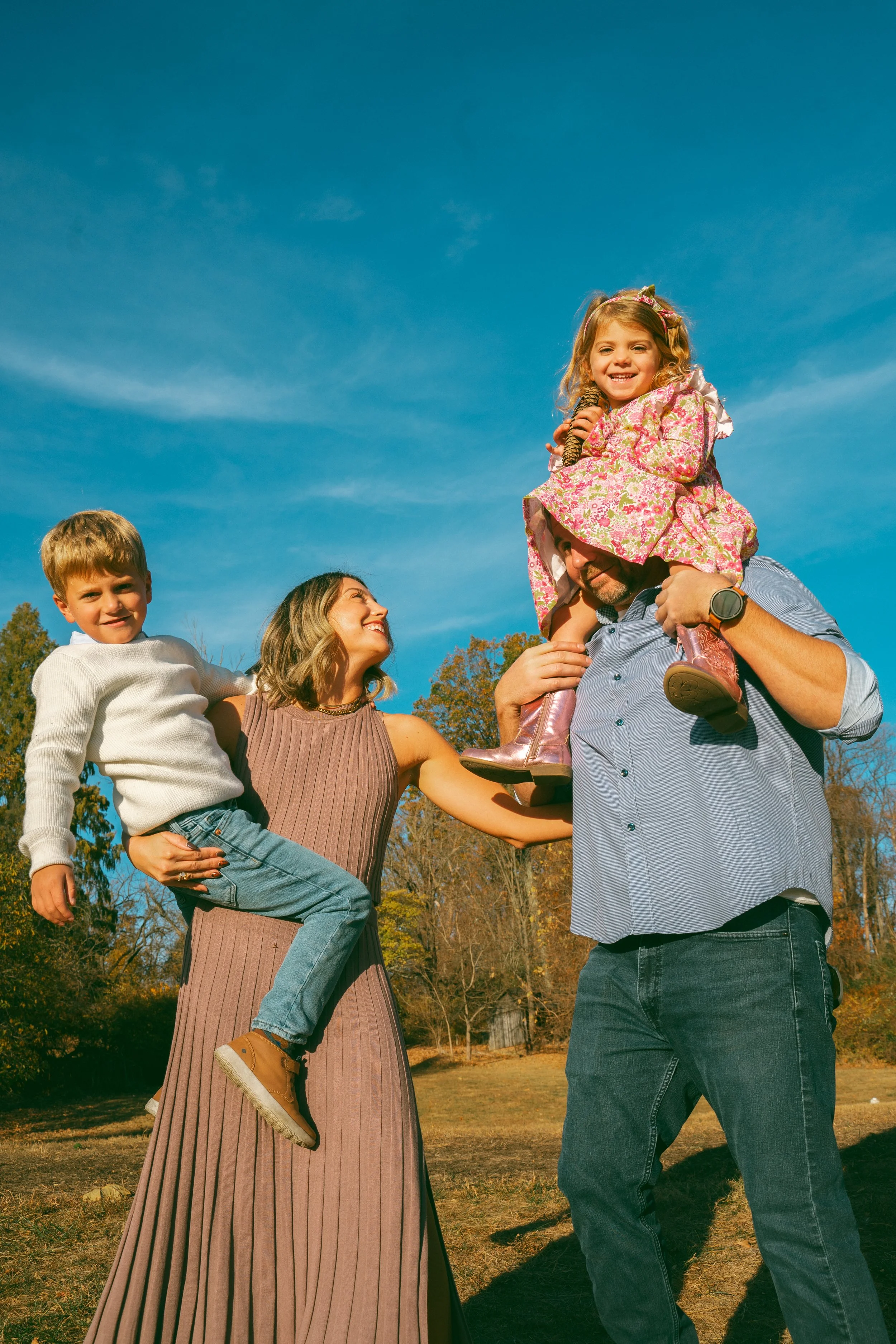 A family enjoying a day outdoors, with a girl sitting on a man's shoulders, a woman smiling, and a young boy standing beside them. The background features trees and a clear blue sky.