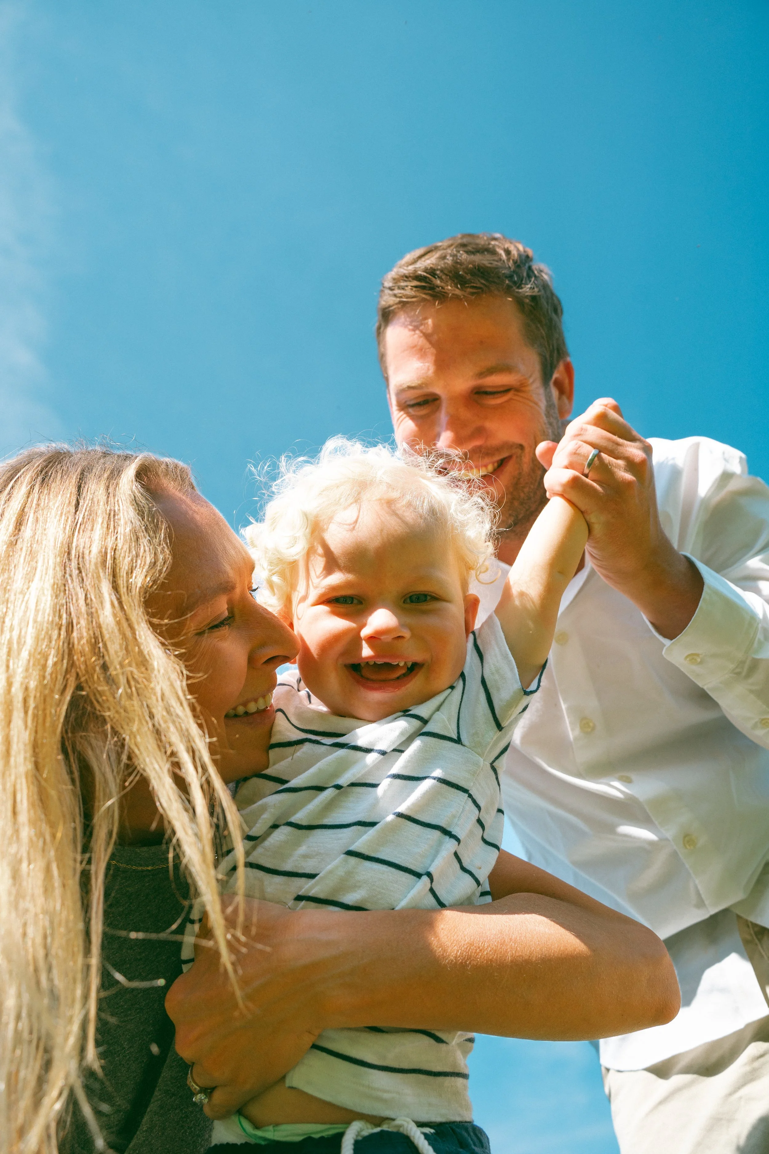 A smiling young boy with blonde curly hair being held up by a woman with long blonde hair, and a man with short brown hair and beard, against a clear blue sky. They seem to be playing or enjoying a moment together outdoors.