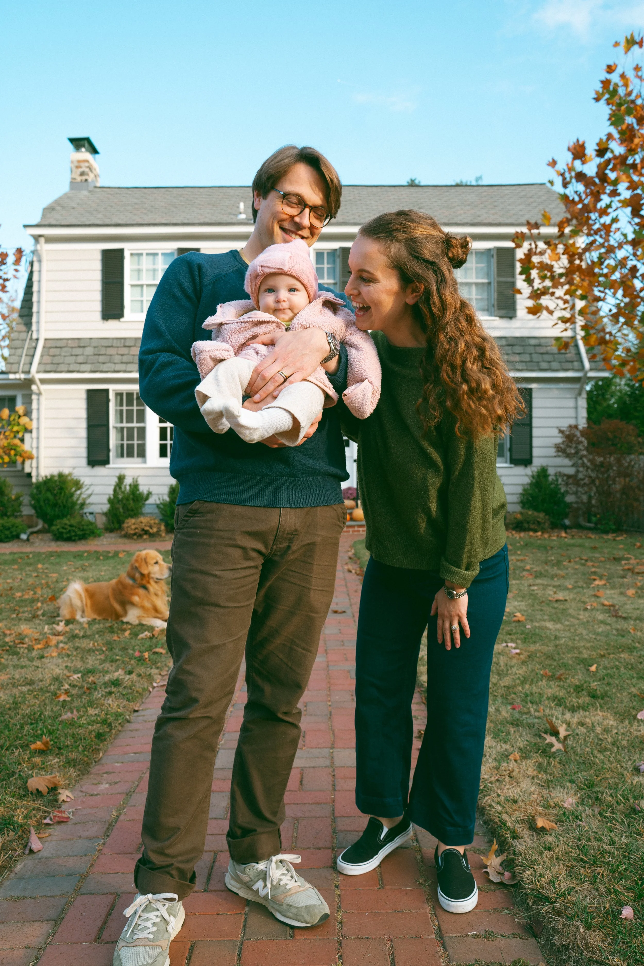 A family with a man, woman, and baby girl standing outside in front of a white house with black shutters. The man is holding the baby, and the woman is leaning toward her. There are trees with autumn leaves and a golden retriever dog lying on the gra