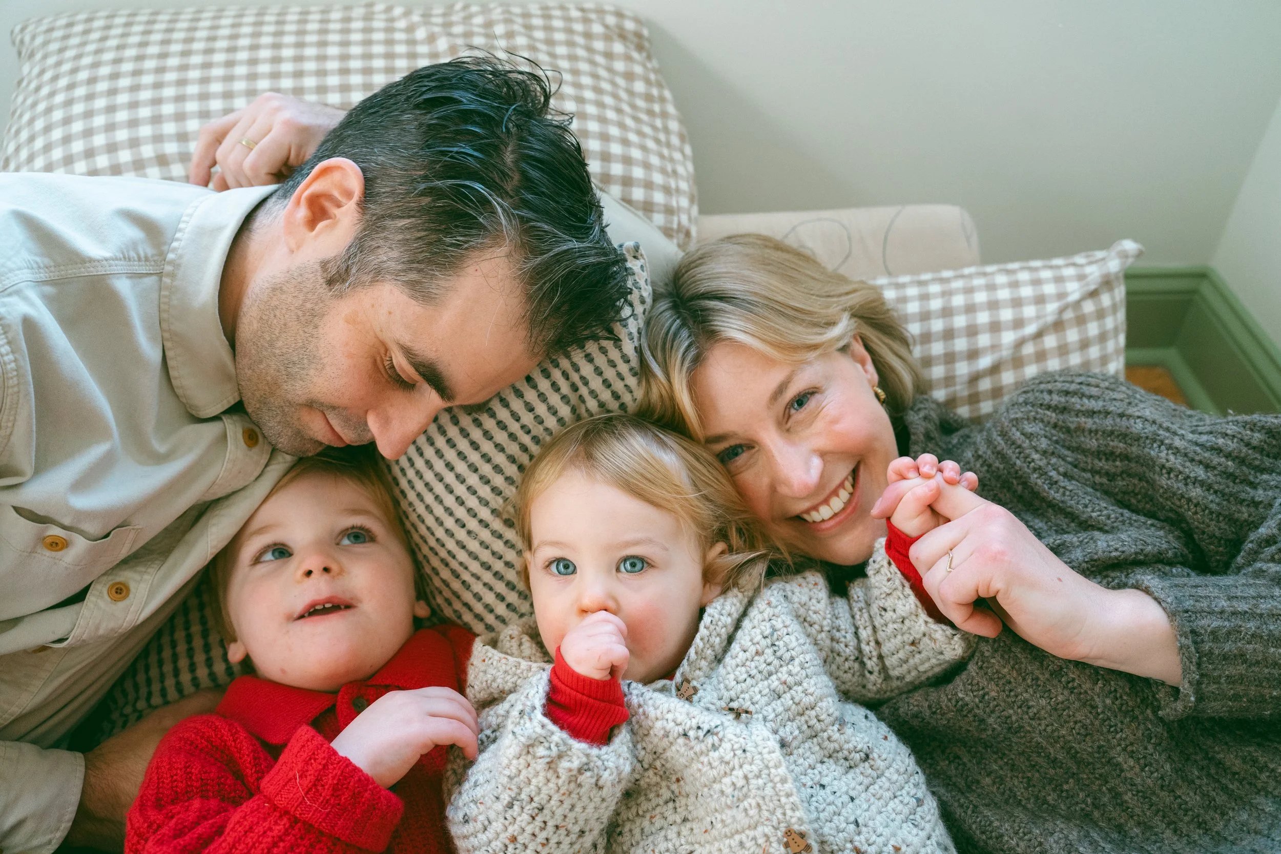 A family of five lying on the bed together, smiling and looking at the camera, with two young children and their parents, all with light skin and blond hair, in a cozy bedroom with checkered pillows and a soft, knitted blanket.