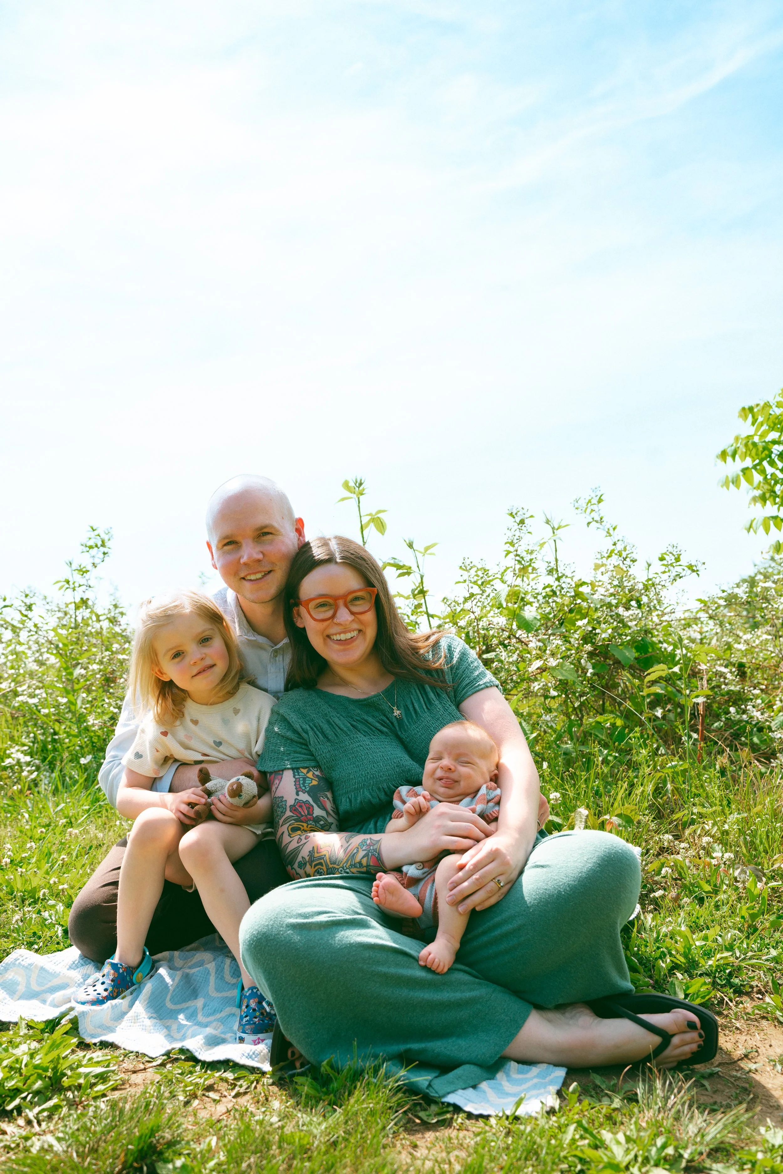 Family of six sitting and lying on a blanket in a green field, surrounded by plants and trees, smiling at the camera under a clear blue sky.