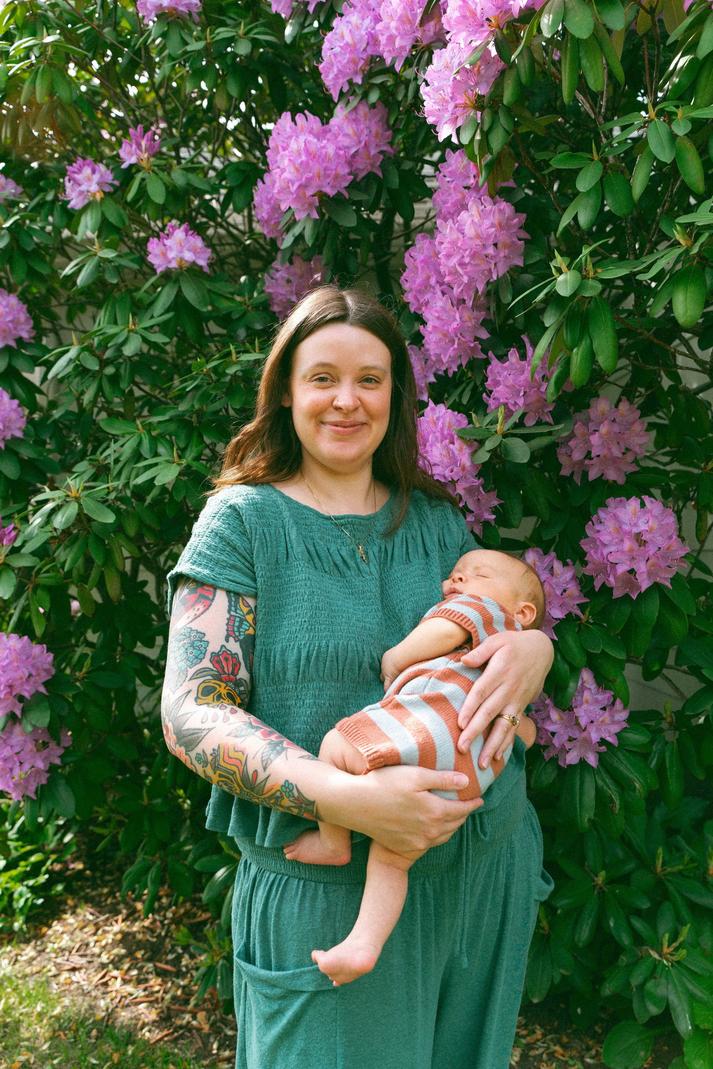 A woman with long dark hair, wearing a green dress and displaying colorful tattoos on her arm, is holding a sleeping baby dressed in striped clothes. They are standing outdoors in front of a large bush with pink and purple flowers.