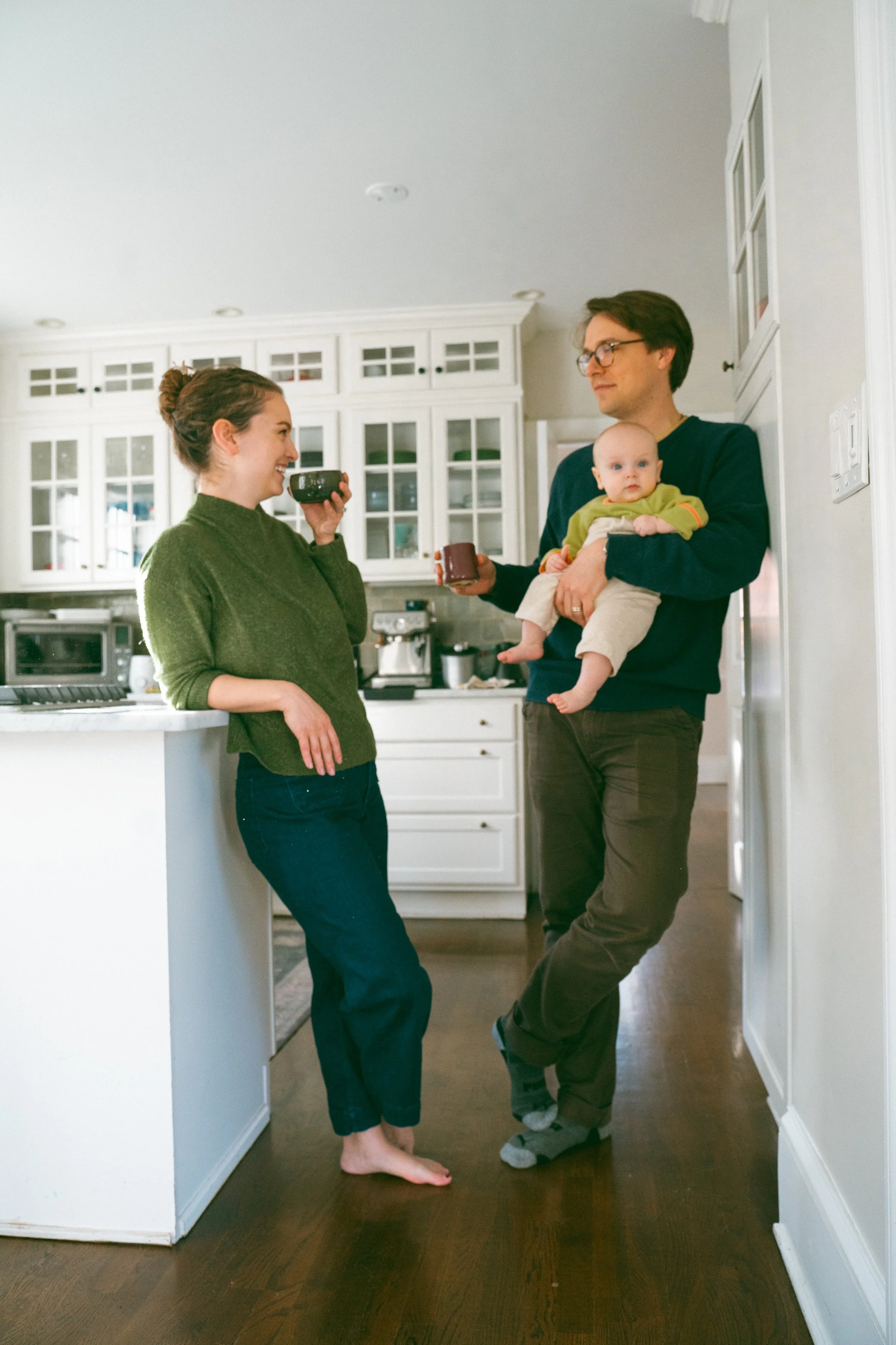 A woman, a man, and a baby are in a kitchen. The woman is smiling and holding a mug, leaning against the counter. The man is holding the baby, who is looking attentively forward. The kitchen has white cabinets and appliances, and a wooden floor.