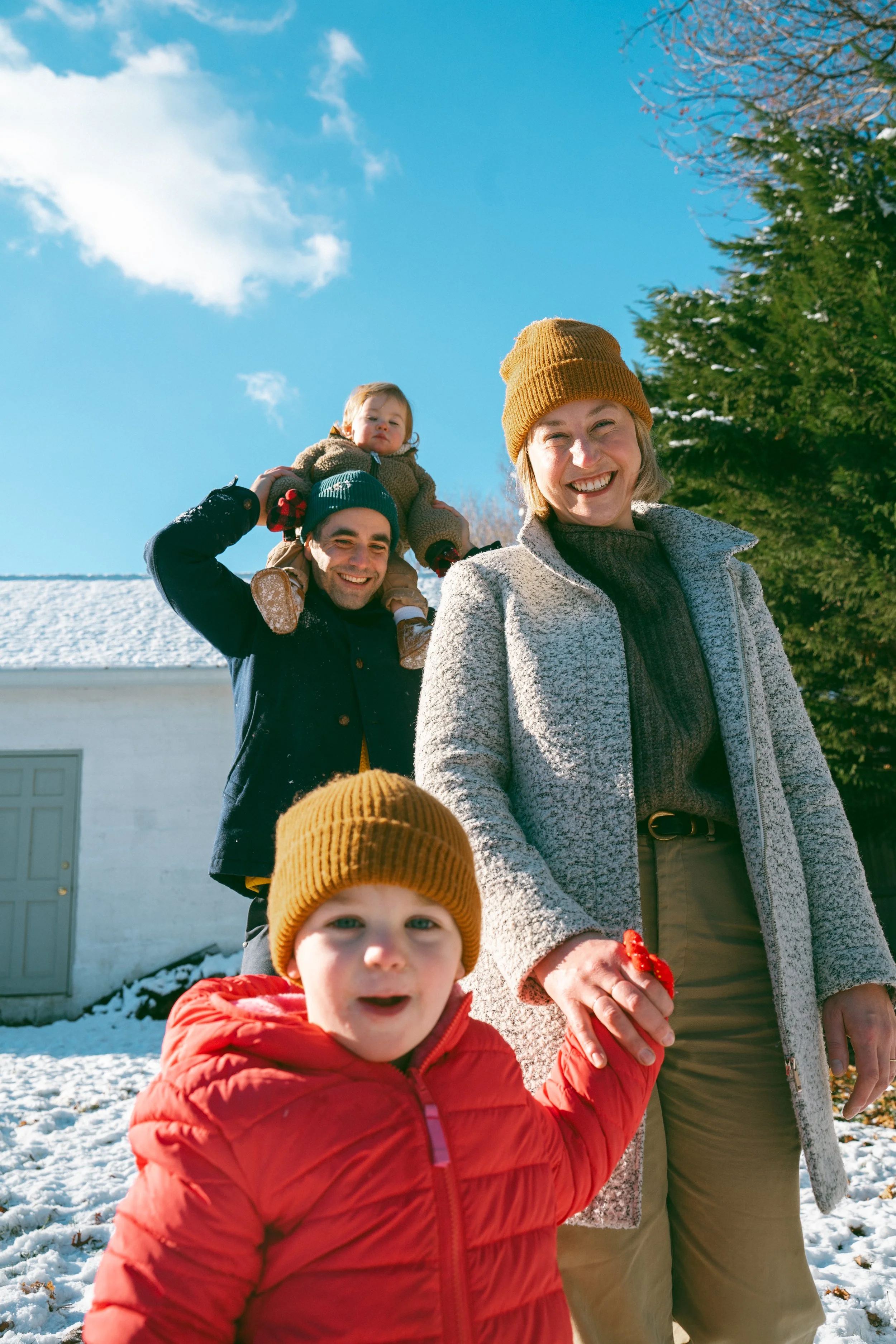 A family outdoors on a snowy day. A woman wearing a brown beanie and cream jacket smiles. A man with a blue beanie carries a young girl on his shoulders. Another child in a red coat and brown hat is in the foreground.