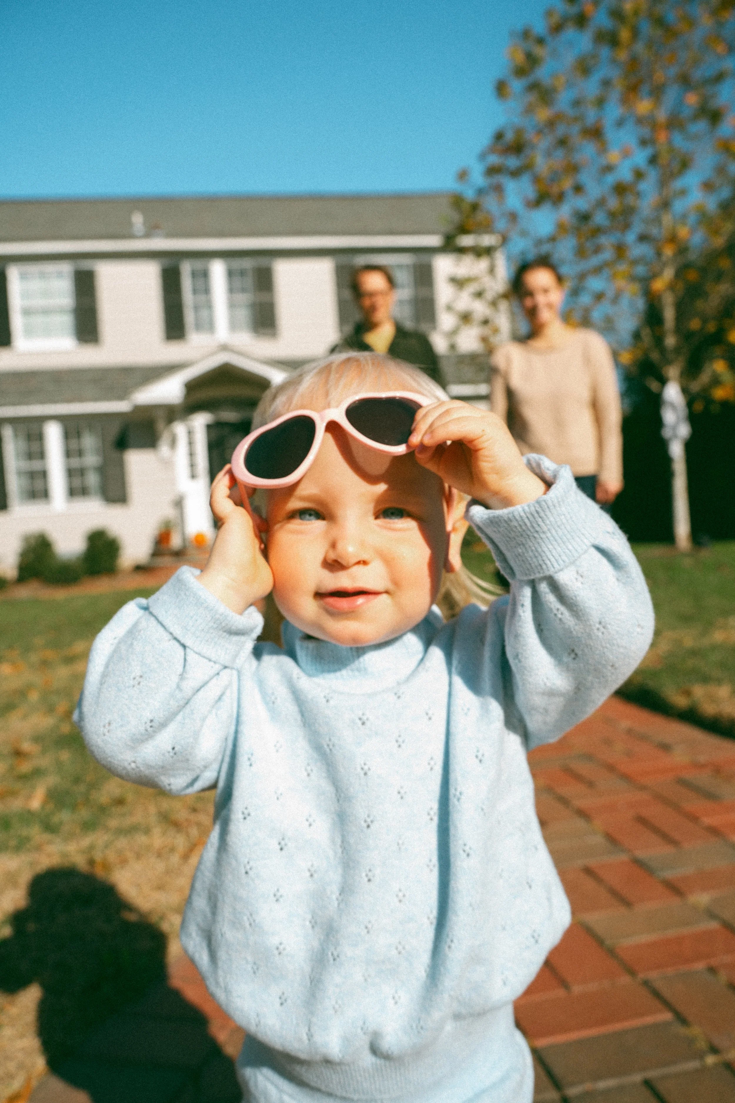 A young child with blonde hair and blue eyes holding pink sunglasses up to their face, outdoors on a sunny day in front of a large house with black shutters, with two women standing in the background near a tree with autumn-colored leaves.