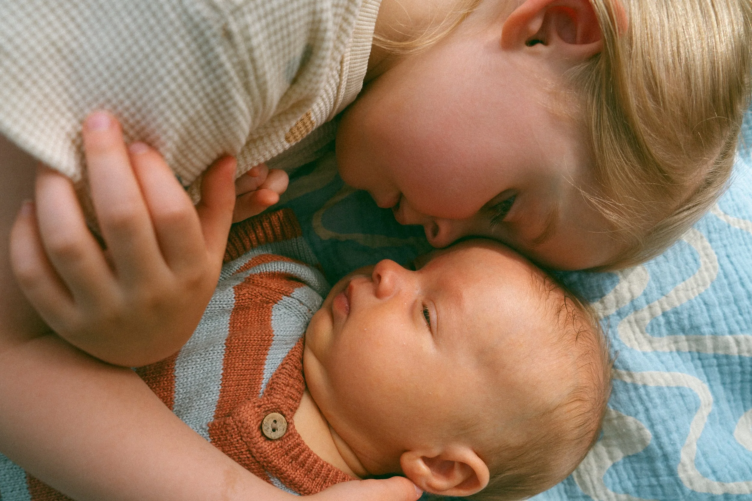 A young child cuddling closely with an infant, both lying on a blanket, sharing a tender moment.