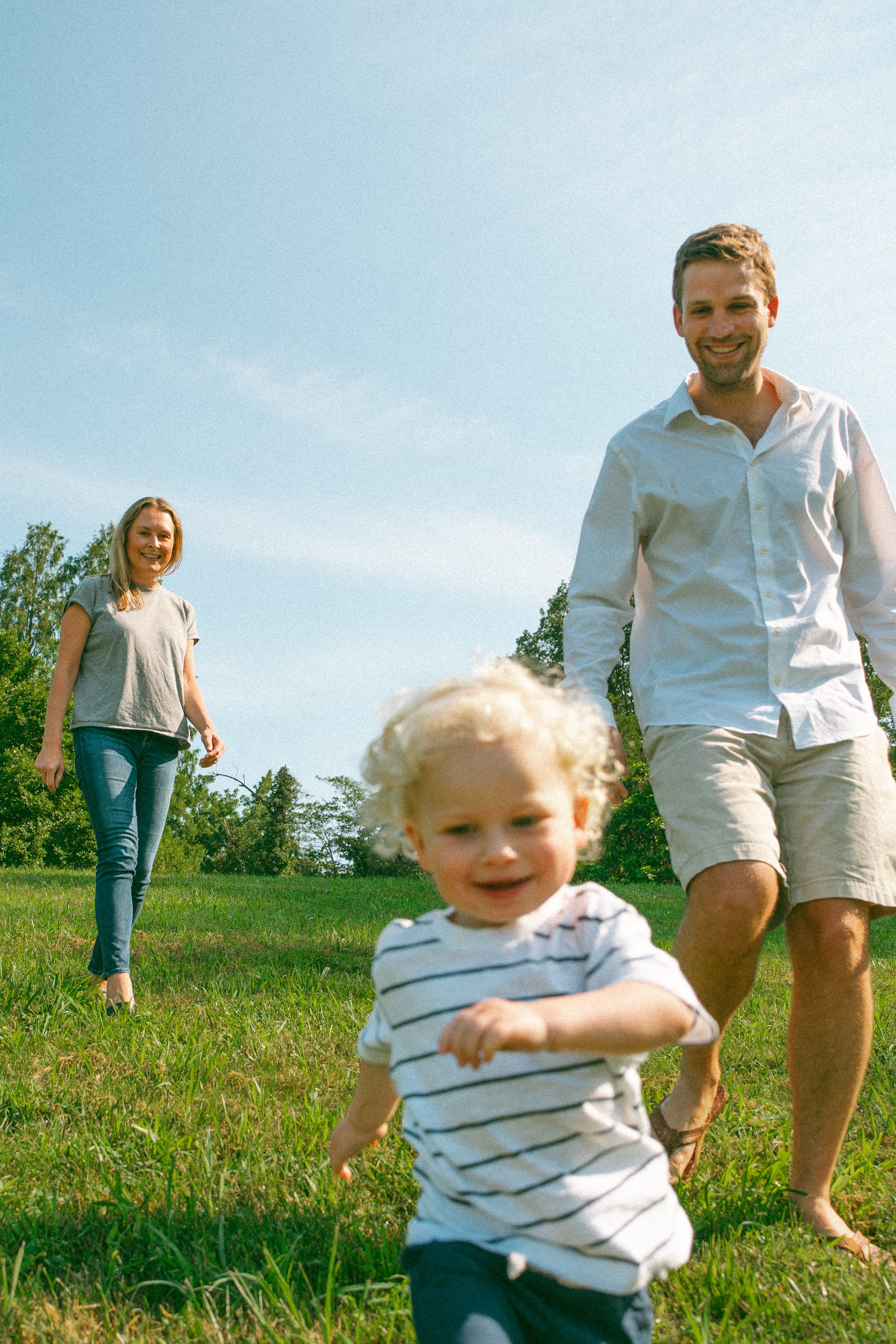 A family enjoying outdoor activities in a grassy field on a sunny day, with a young child in the foreground running towards the camera and smiling, and two adults smiling and walking behind him.