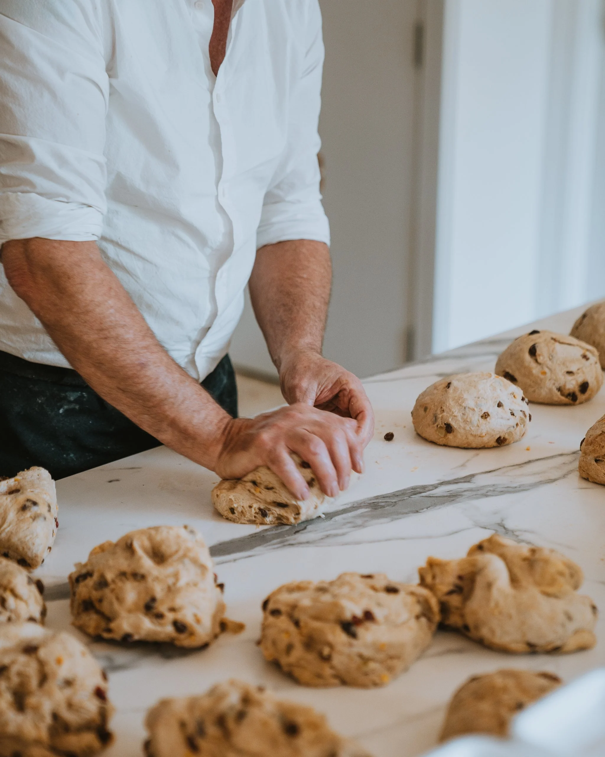 A person wearing a white shirt is kneading dough with chocolate chips on a marble surface, surrounded by prepared chocolate chip cookie dough balls.