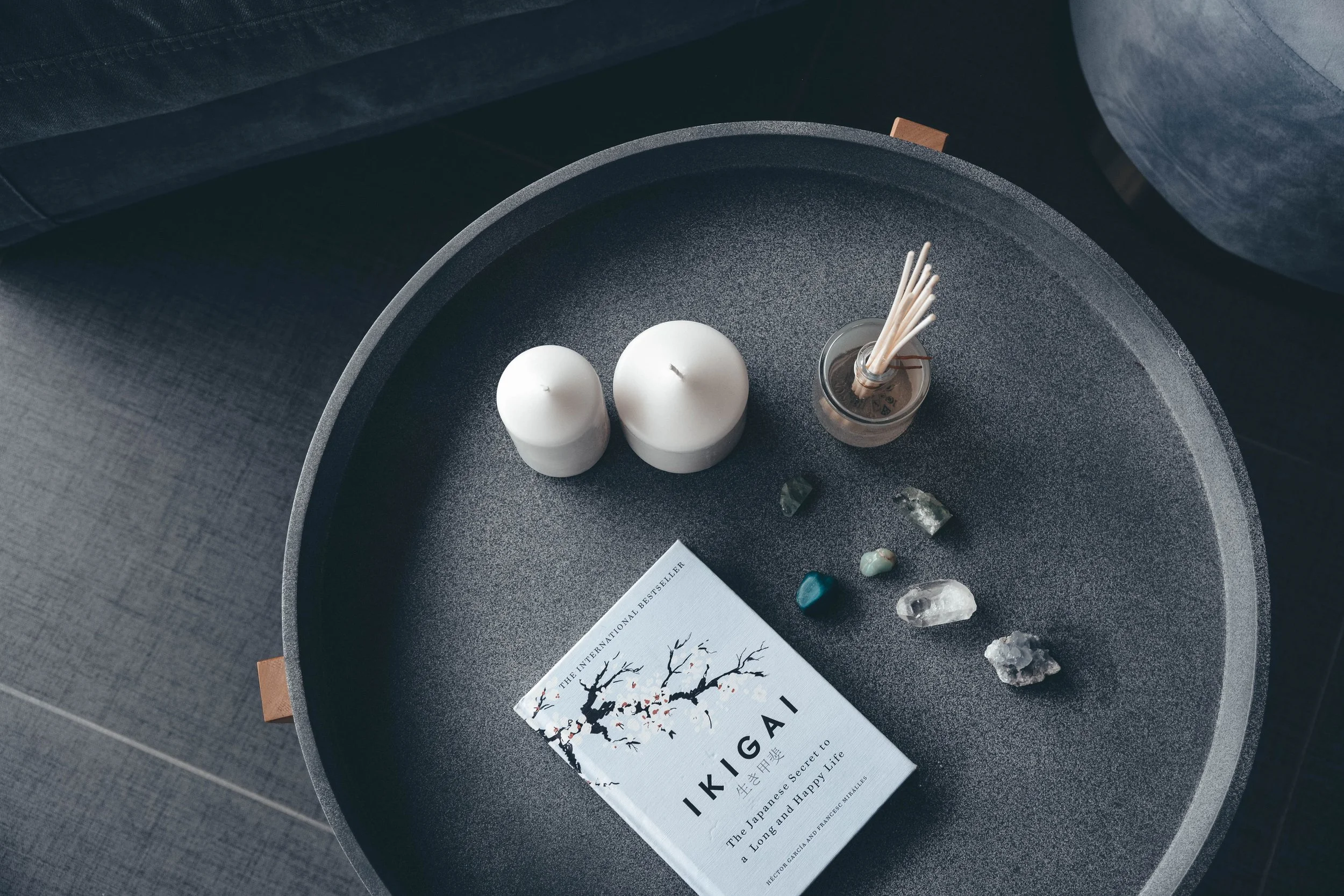 A round black table with two white candles, a small container with incense sticks, a hand-shaped item, and a book titled 'Ikigai' on top of a brown notebook.