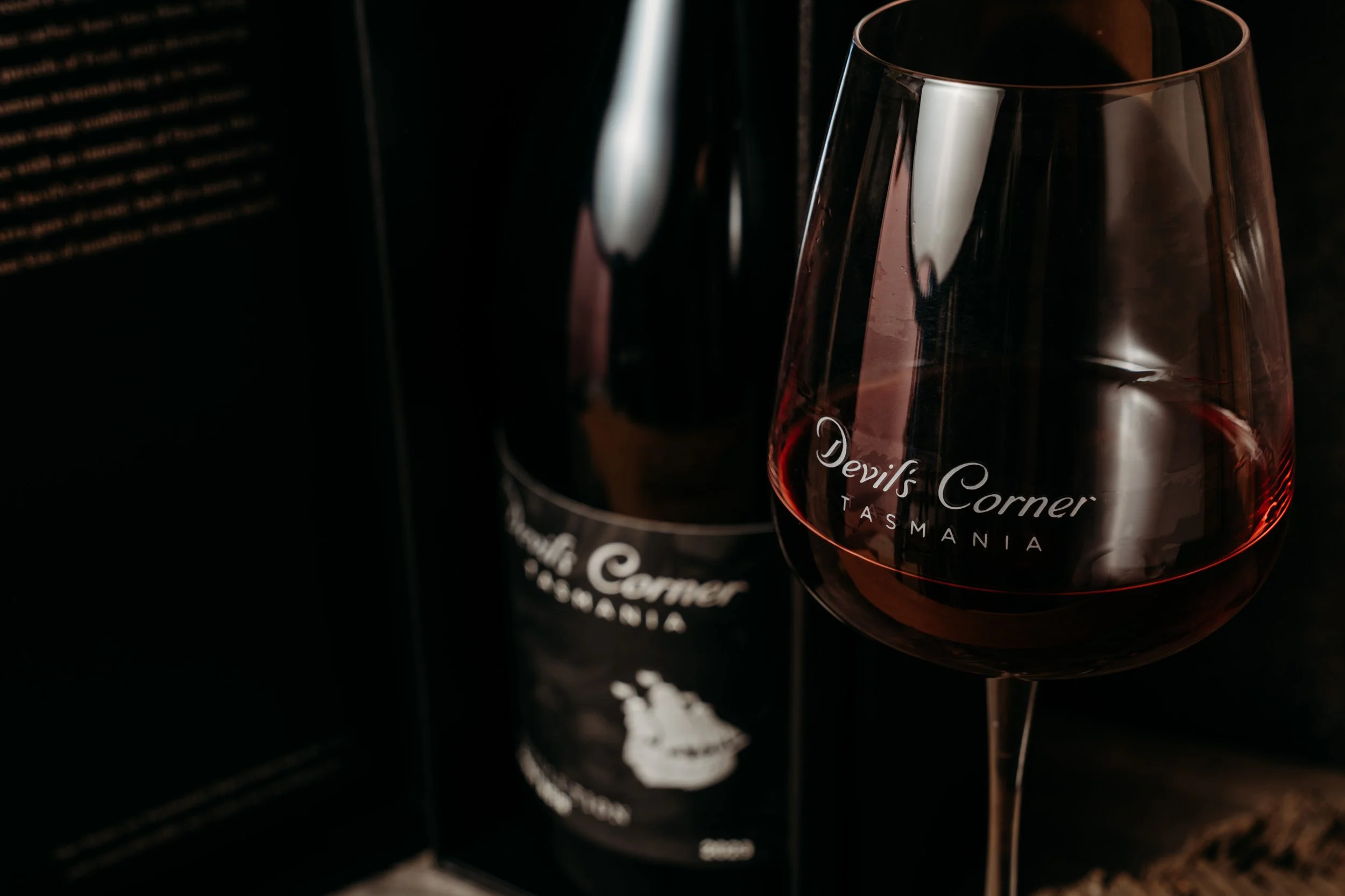 A glass of red wine with the label 'Devil's Corner Tasmania' in front of a wine bottle with the same label, on a dark background.