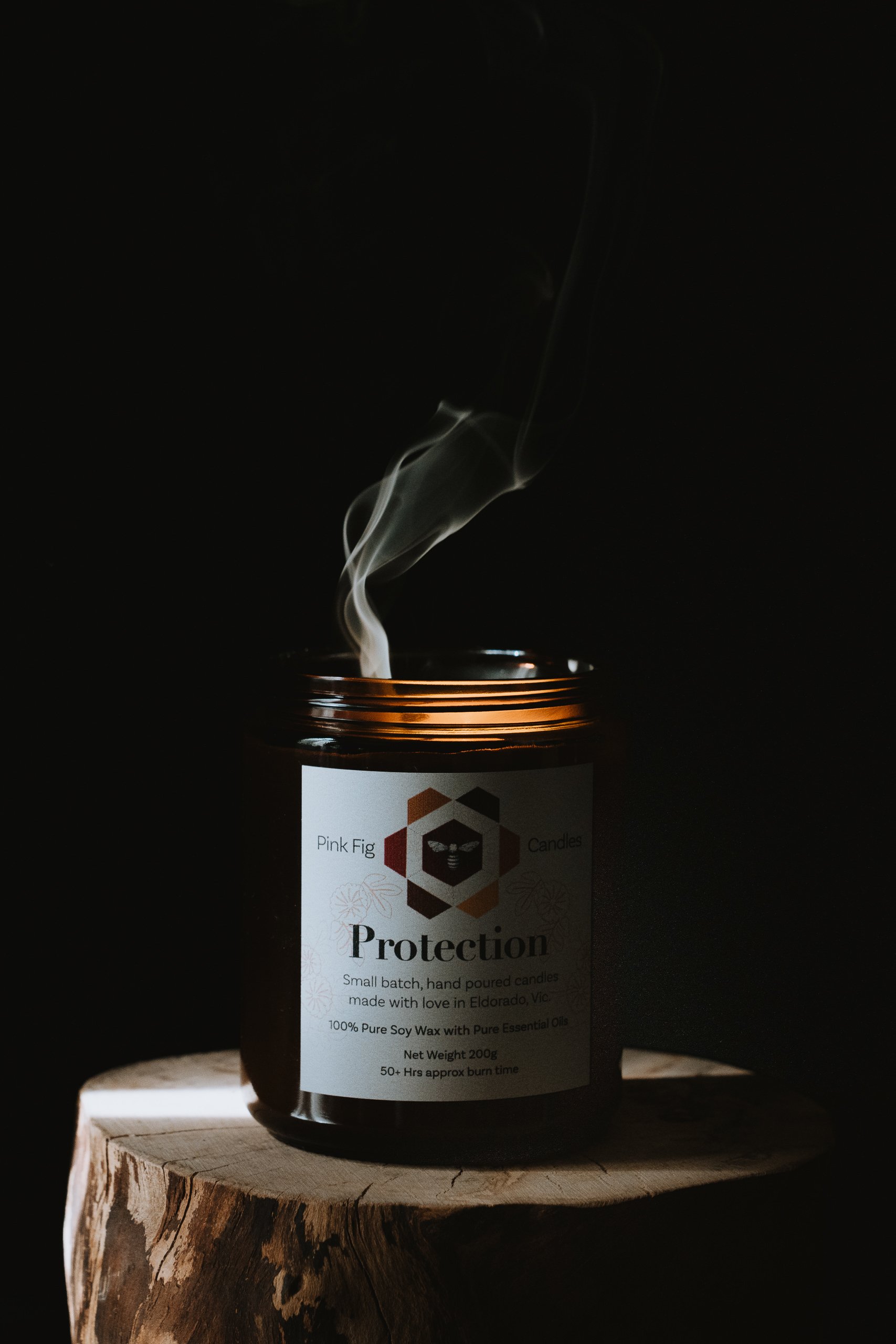 A black candle jar with a white label that reads 'Protection' and features a honeycomb pattern logo, placed on a wooden surface with a small amount of smoke rising from the unburned wick. The background is dark, highlighting the candle and smoke.