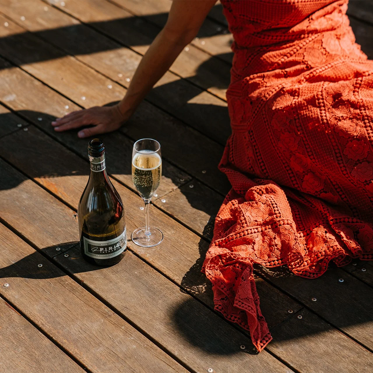 A person in a red lace dress sitting on a wooden deck, with a bottle of champagne and a glass of champagne nearby.