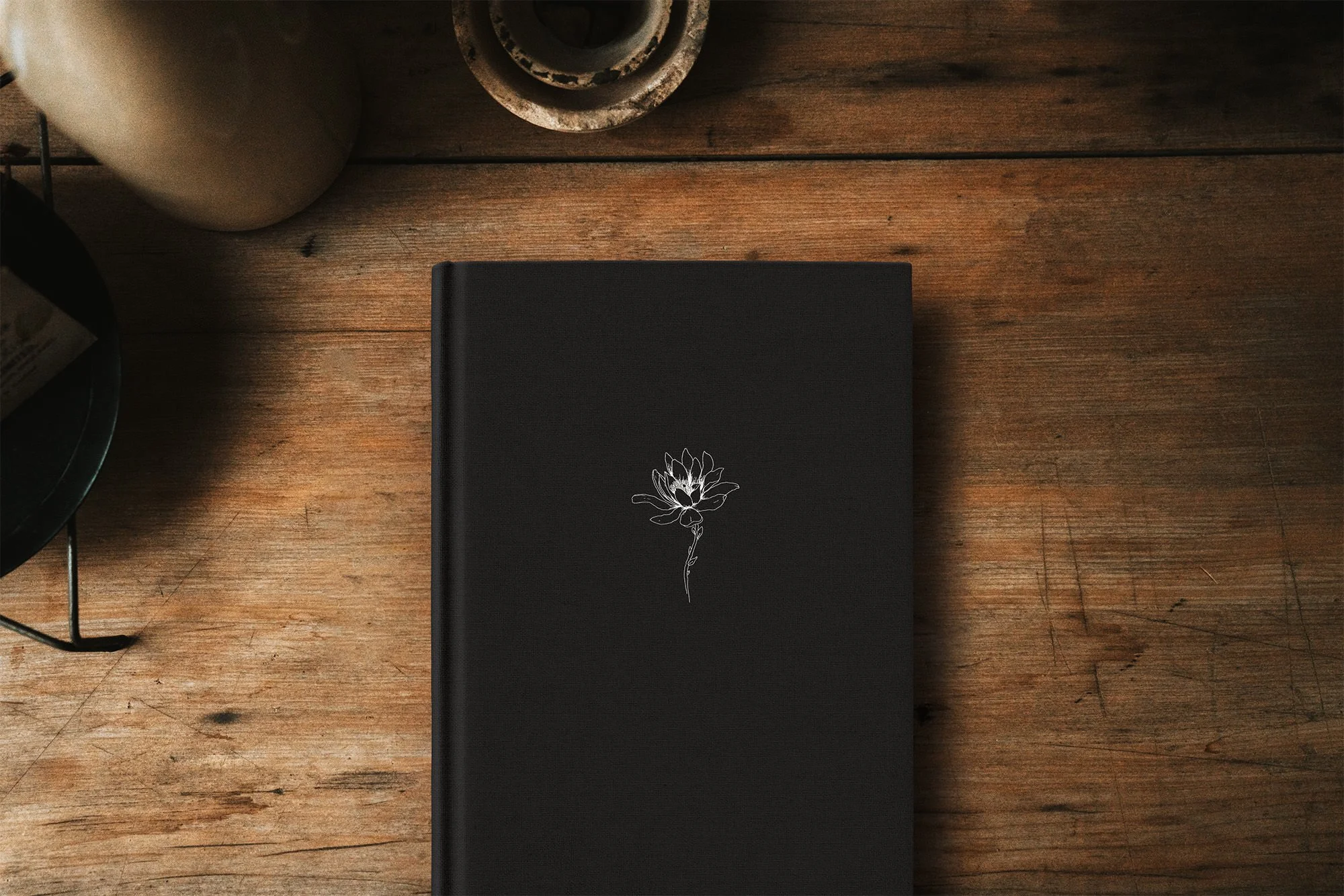 Black notebook with a white floral illustration on the cover, placed on a wooden surface with a coffee cup and a rustic coaster nearby.