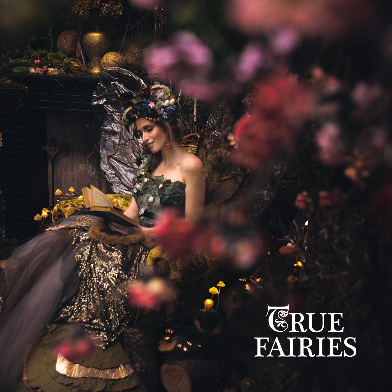 A woman dressed as a fairy sitting in a whimsically decorated garden, reading a book surrounded by pink blossoms and decorated with fairy wings and a floral crown. The image has the words 'True Fairies' in the bottom right corner.