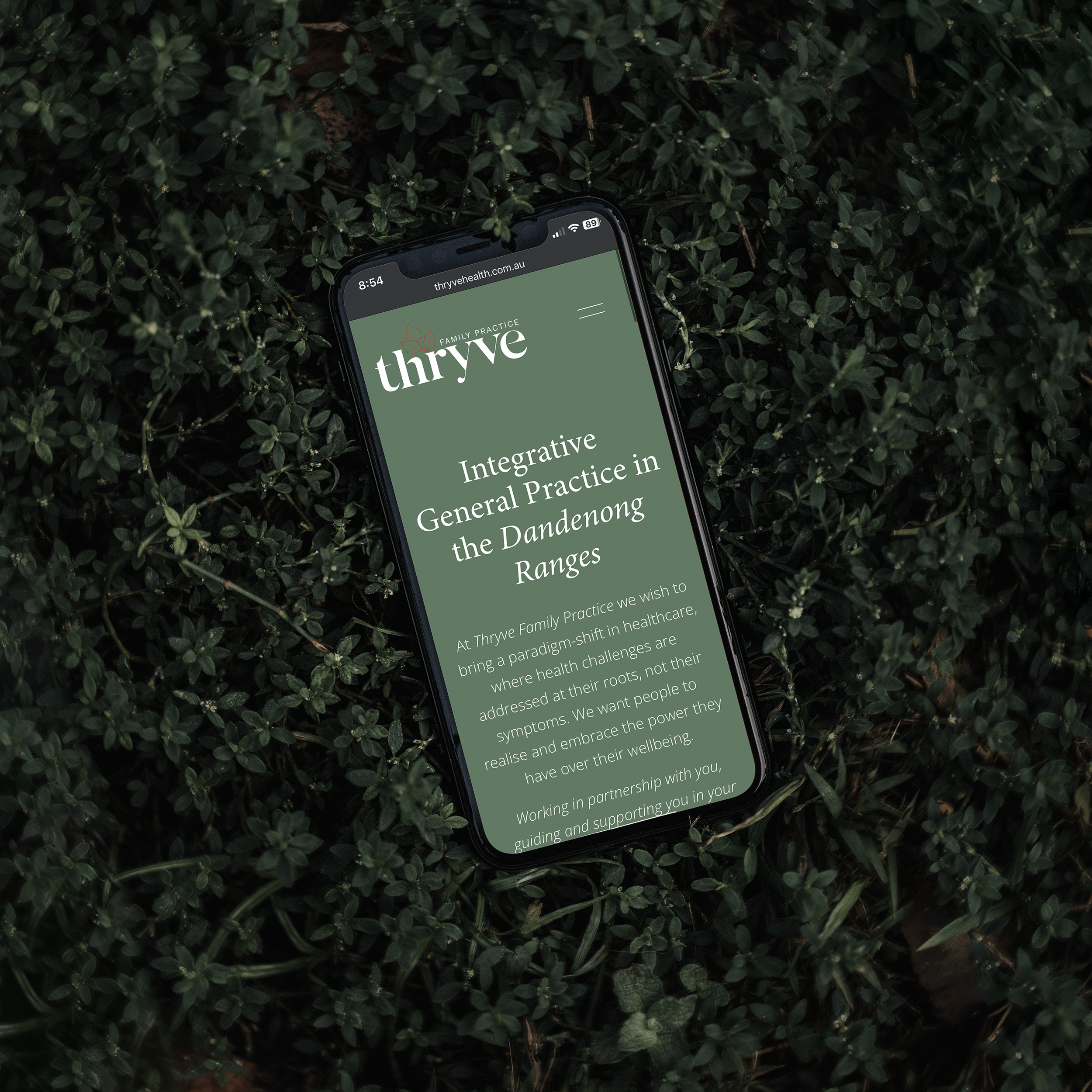 A smartphone lying on a bed of green plants displaying a webpage from Thrive Family Practice about integrative healthcare in the Dandenong Ranges.
