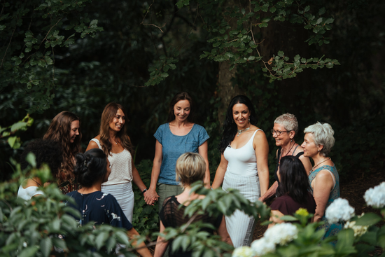 Group of women standing outdoors among green trees and plants, smiling and engaging in conversation.