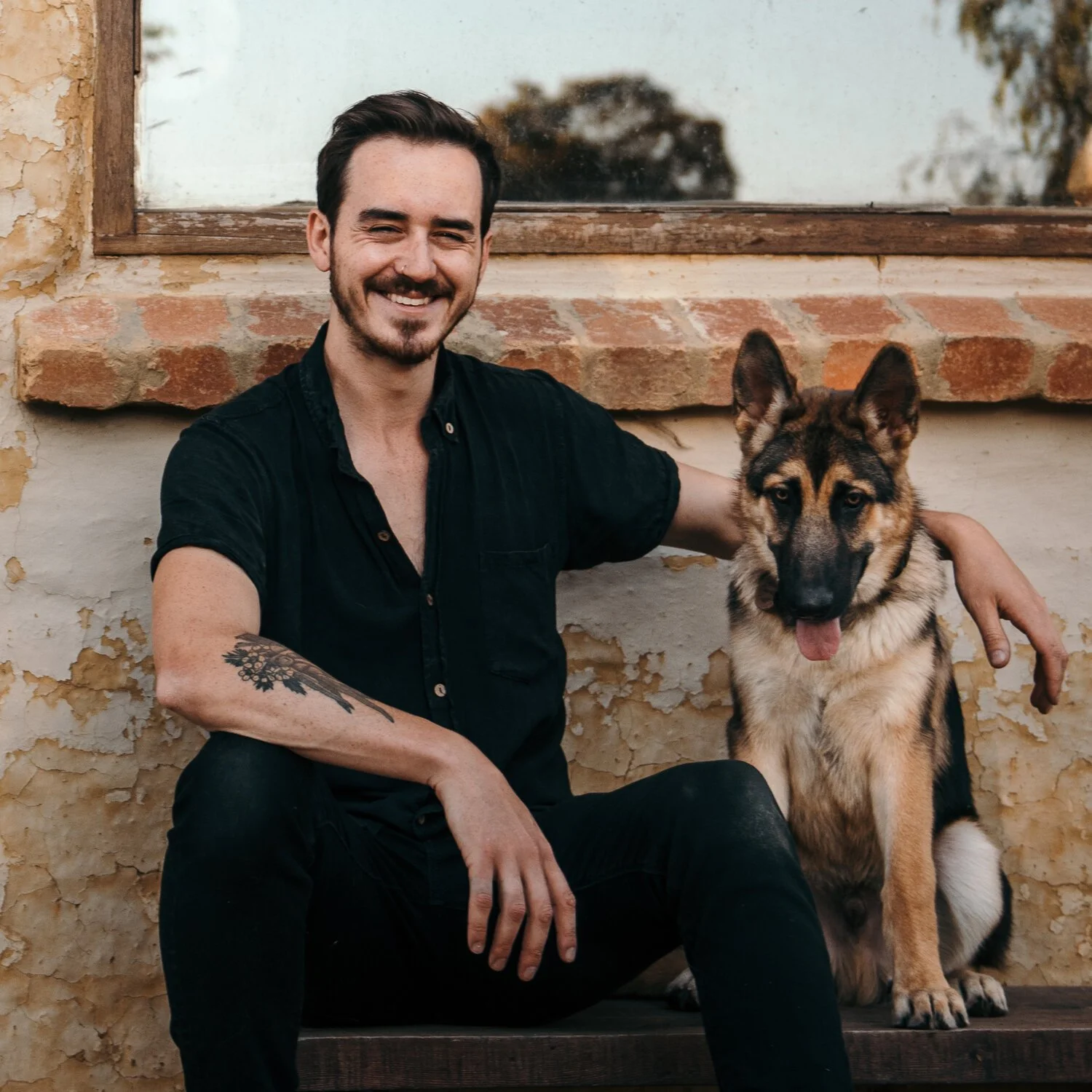 A smiling man with dark hair and a beard sitting next to a German Shepherd dog against a weathered wall with a window.
