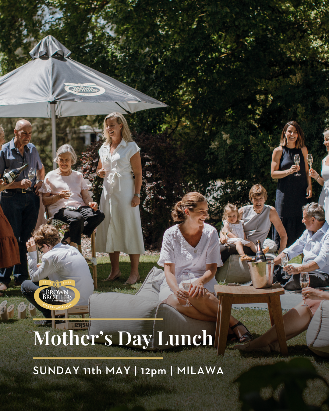 Group of people celebrating Mother's Day lunch outdoors under a large patio umbrella, with women holding glasses of champagne, smiling and chatting, surrounded by greenery and casual seating.