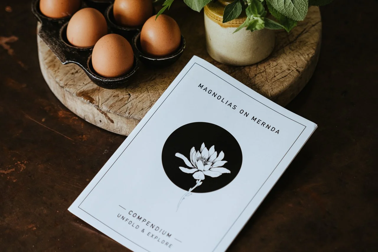 A menu or brochure titled 'Magnolias on Mernda' with a black circle featuring a white magnolia flower illustration, placed on a dark wooden surface next to a wooden board holding eggs and a small plant.
