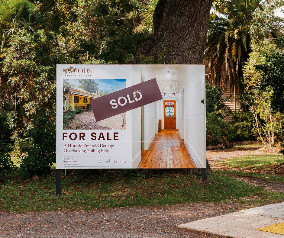 Real estate for sale sign with a 'SOLD' label on it, advertising a historic cottage in a wooded area.