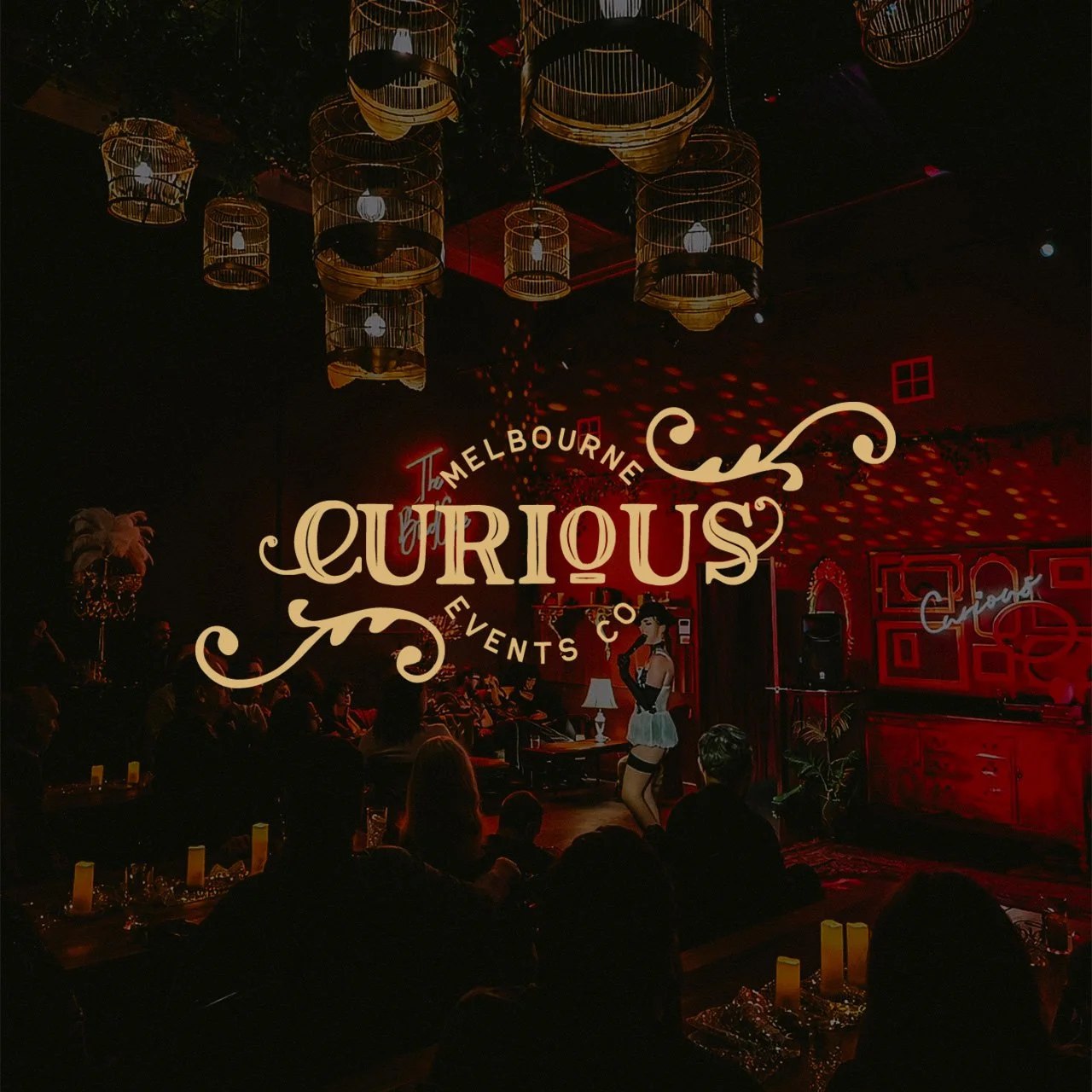 Dimly lit bar or lounge with a stage where a performer dressed in a red and blue outfit is performing. Warm lighting from overhead lamps and candles on tables. Audience seated around tables watching the performance. Decor includes framed art and neon signs, with a large logo for 'Melbourne Curious Events' overlaying the image.