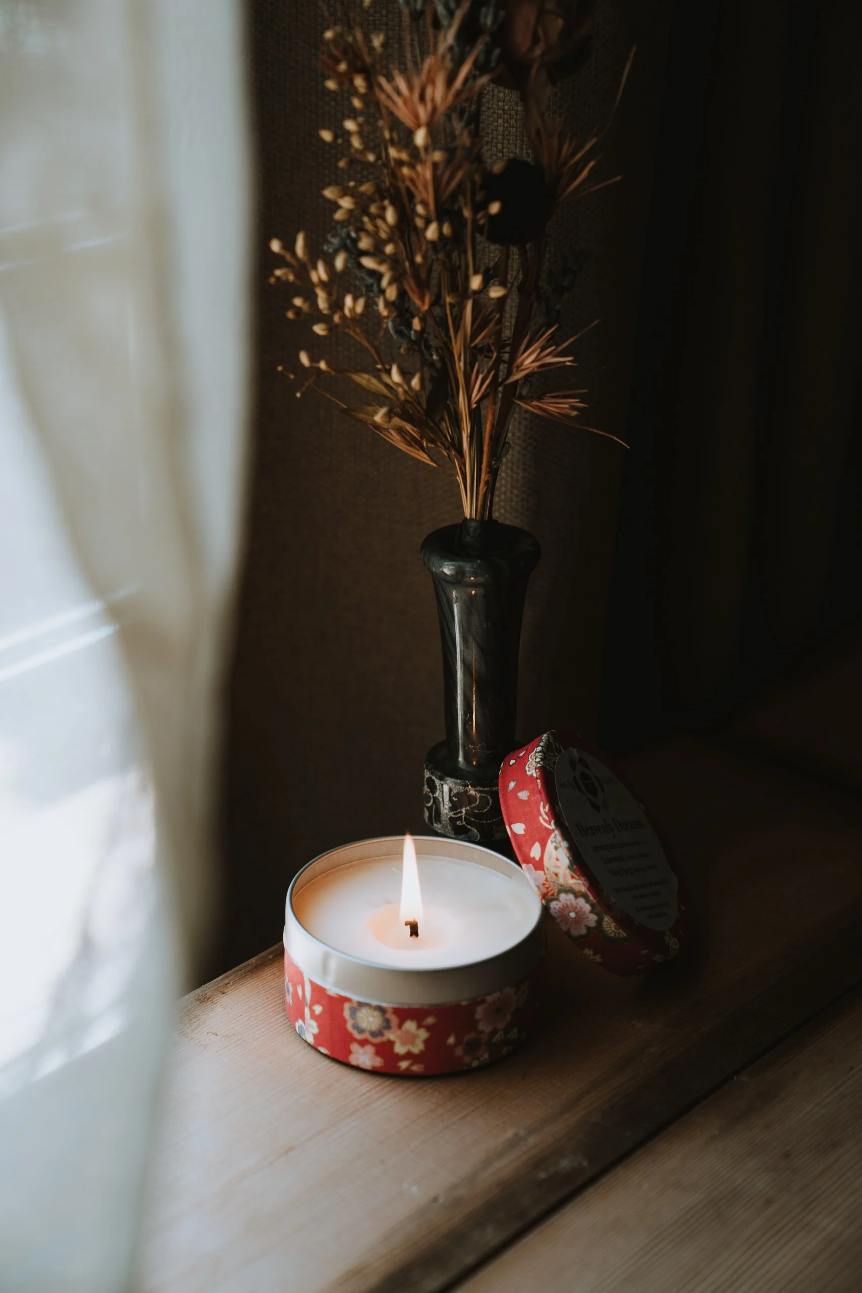 A lit white candle inside a floral-patterned candle holder on a wooden surface. Behind it, there's a black vase containing a dried flower arrangement with small white flowers and long, thin, reddish-brown leaves. Part of a red and white floral patterned object or gift wrap is visible to the right of the candle.