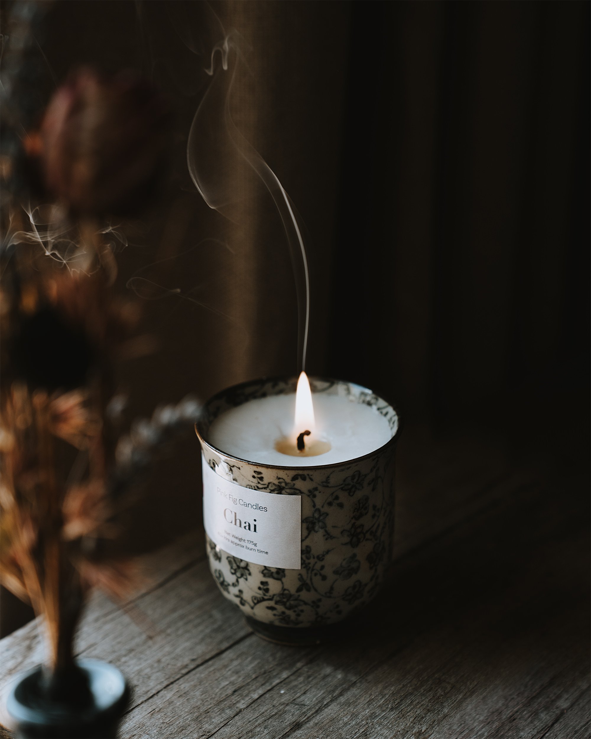 A lit candle with a decorative black and white patterned holder on a wooden surface, with smoke rising from the wick.