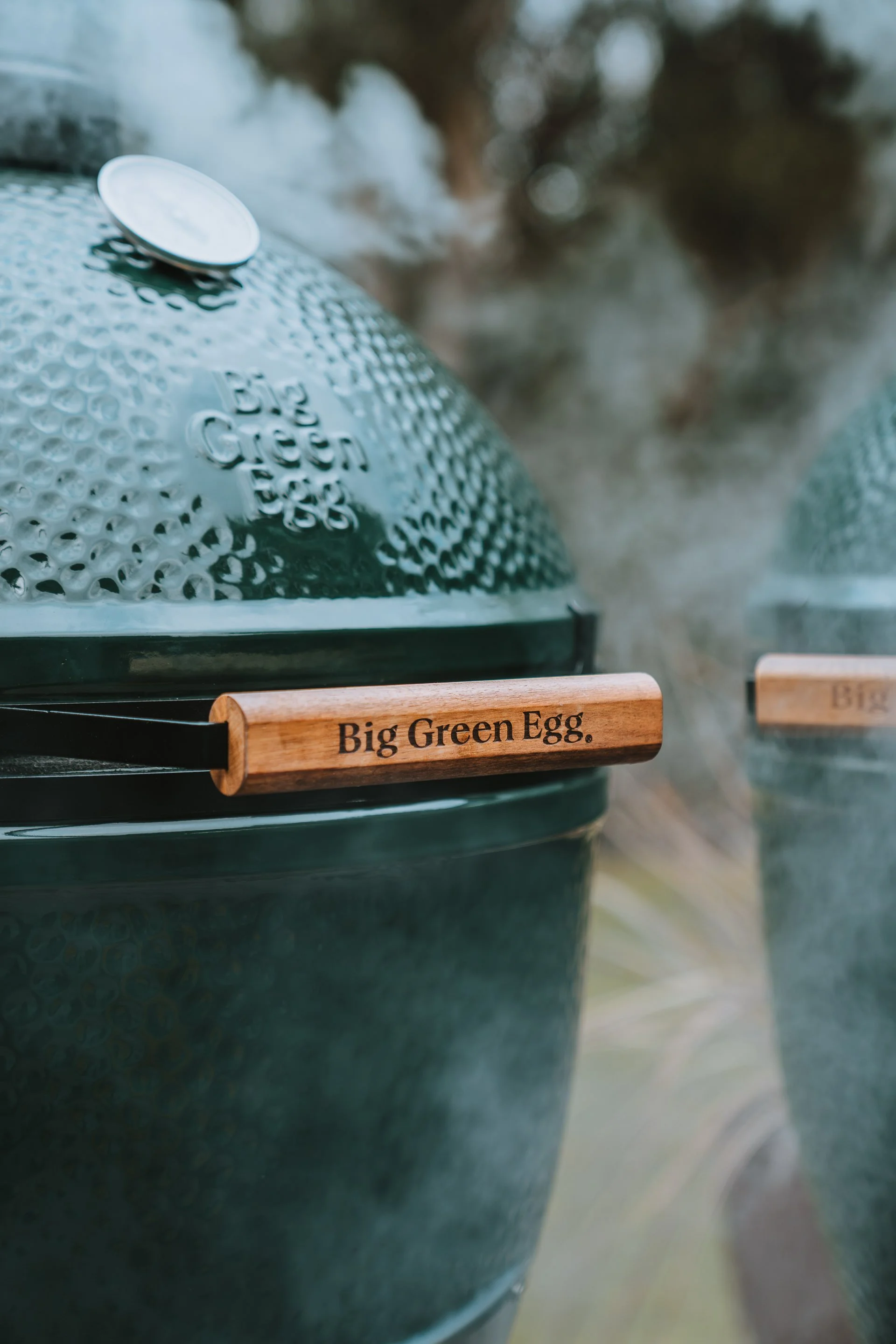 Close-up of a green ceramic grill with a wooden handle labeled 'Big Green Egg', with smoke or steam rising from the background.