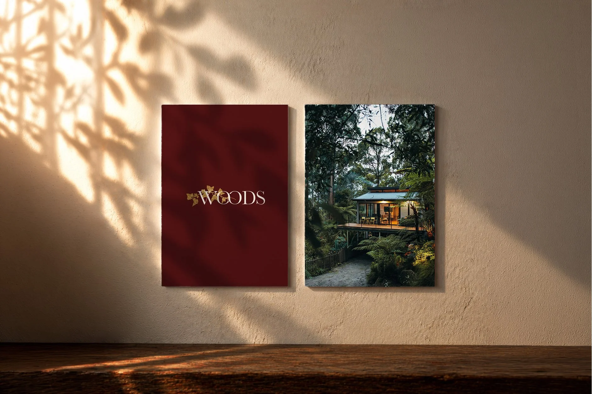 Two framed posters on a beige wall are illuminated by warm light. The left poster is deep red with the word 'WOODS' in white and gold, while the right poster depicts a house surrounded by trees at dusk.
