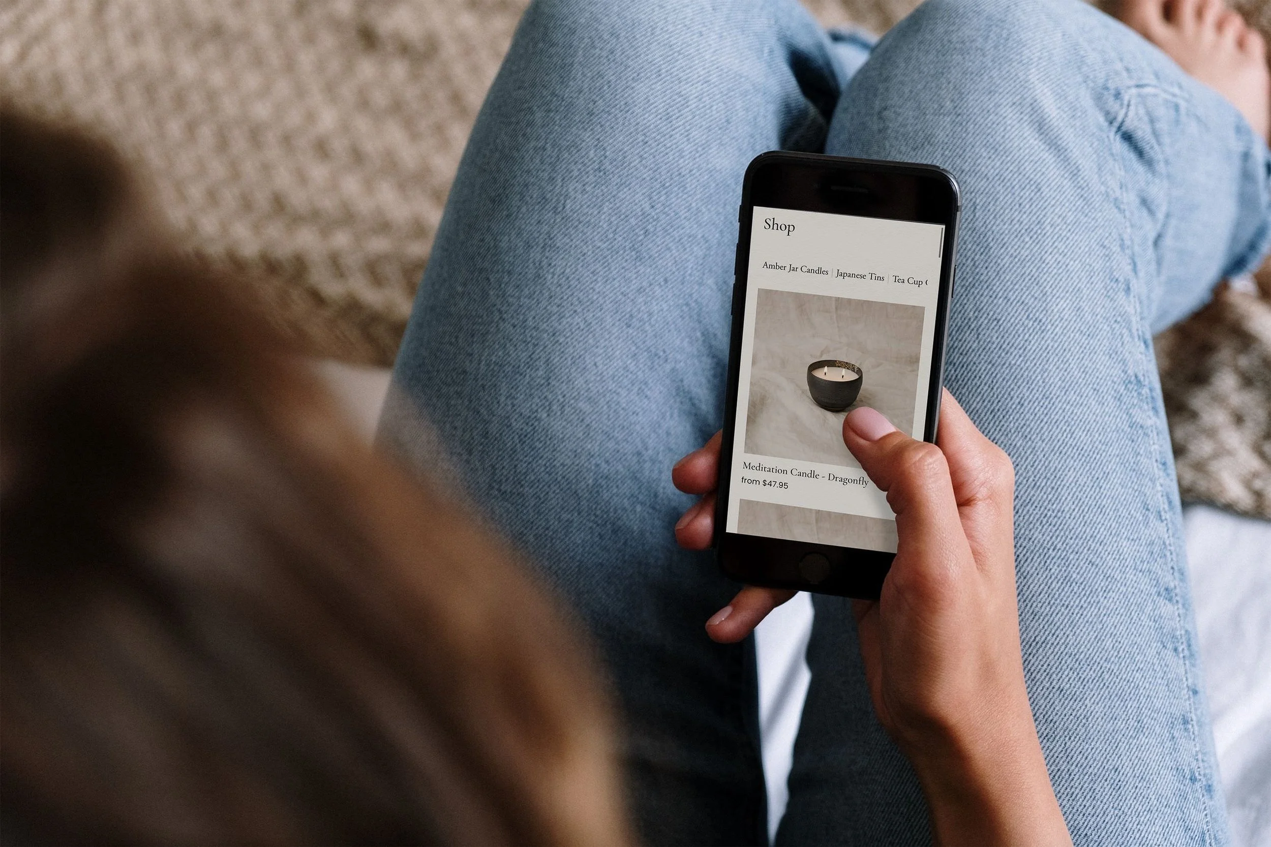 A person sitting on a beige carpeted floor, wearing blue jeans, holding a smartphone displaying an online shop for candles, including amber jar candles, Japanese tins, and tea cups.