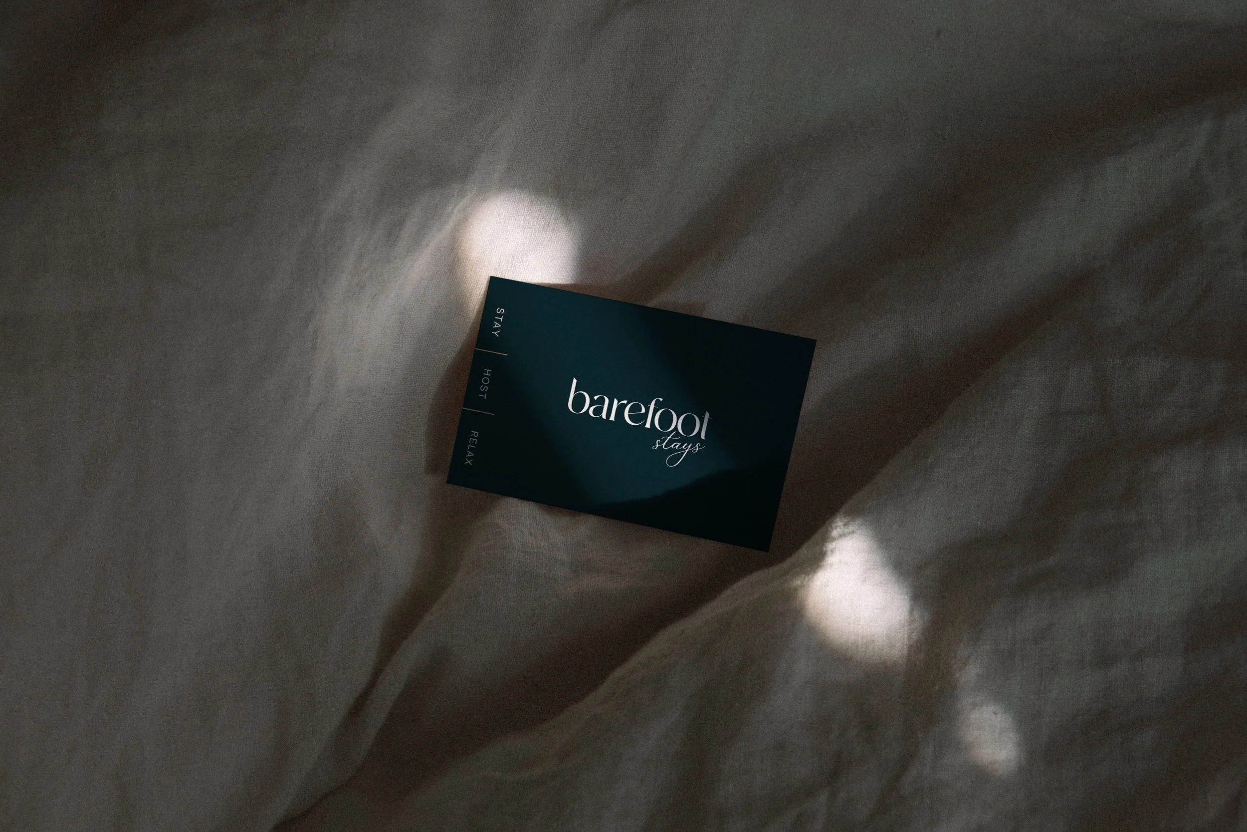 A black card with the words "barefoot stays" written on it, lying on a crumpled light-colored fabric surface.