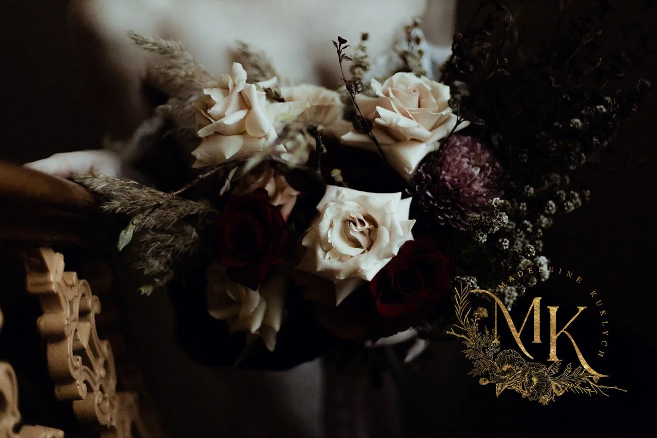 A dark bouquet of flowers with white and dark red roses, dried and purple flowers, and greenery, with a gold monogram and floral logo reading 'Madeleine Kuklych' in the bottom right corner.
