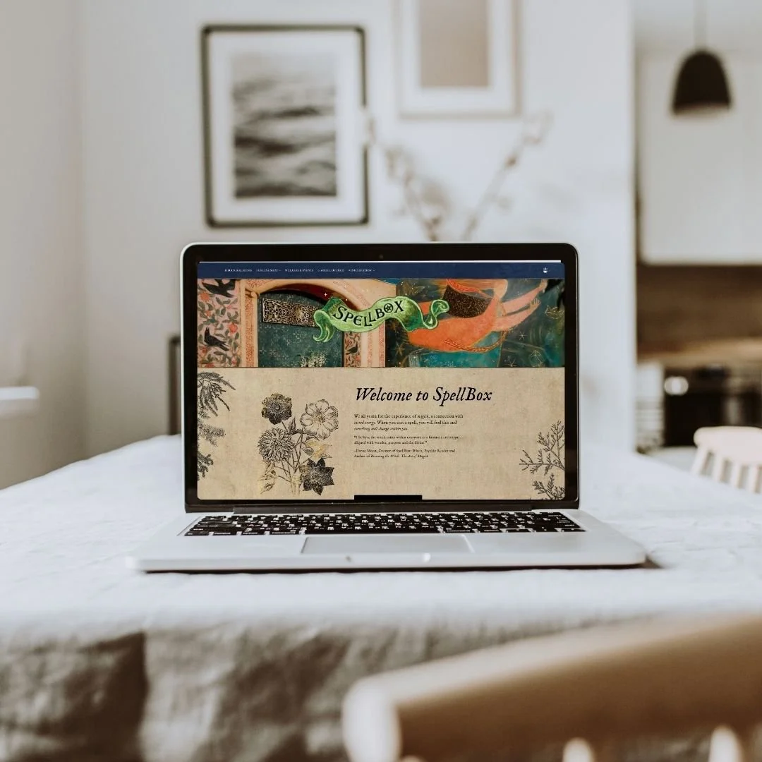 Laptop on a dining table displaying the website 'SpellBox' with decorative botanical illustrations on a beige background.