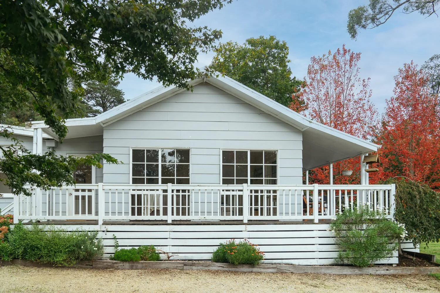 White house with a front porch, surrounded by trees with autumn foliage, and a small garden in front.