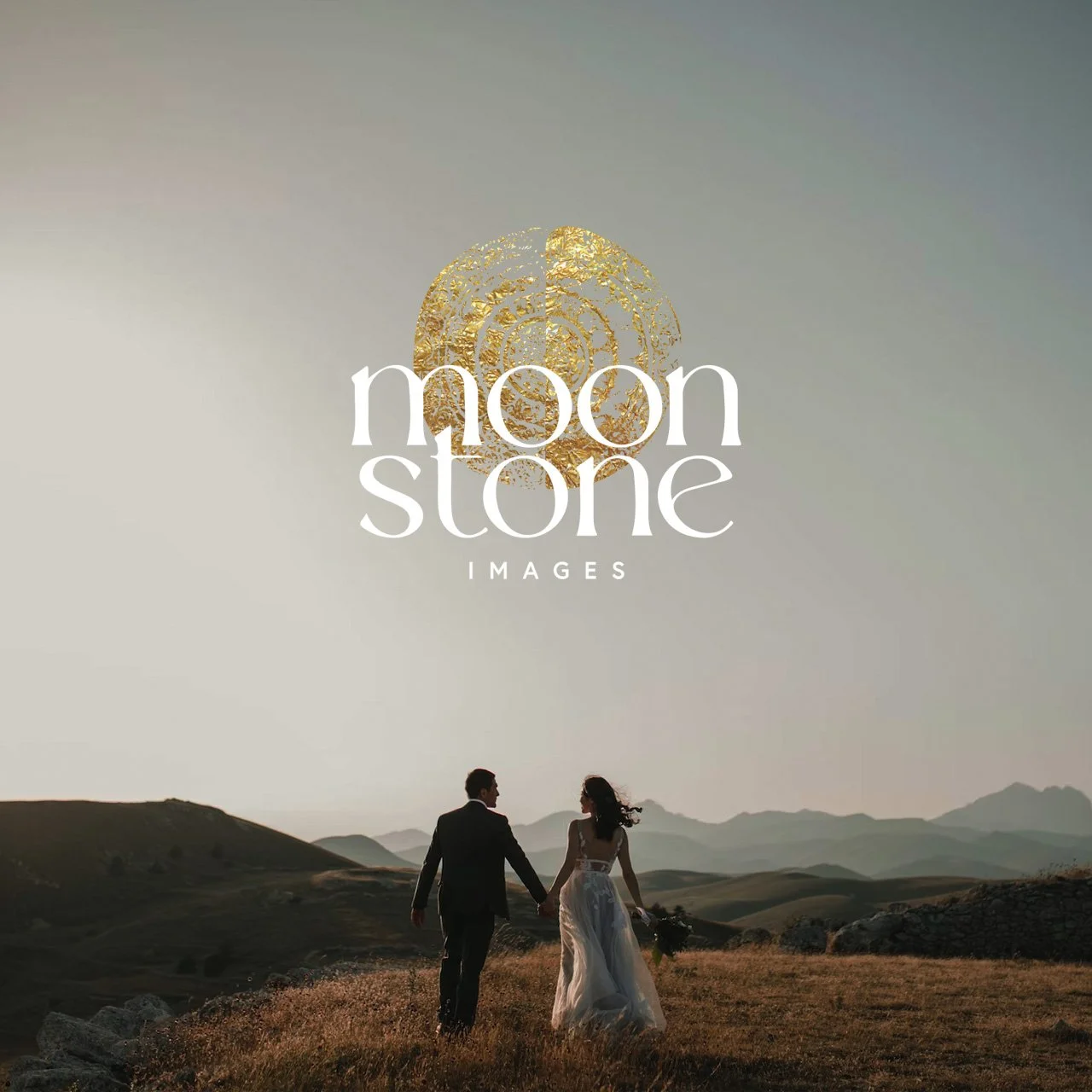 A couple in wedding attire holding hands and walking across a grassy field in a mountainous landscape during sunset or sunrise.